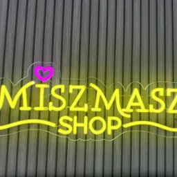 Neonowy szyld 'MISZMASZ SHOP' z różowym sercem, umieszczony na tle pionowych, ciemnych paneli ściennych. Żółte światło neonu oświetla napis.