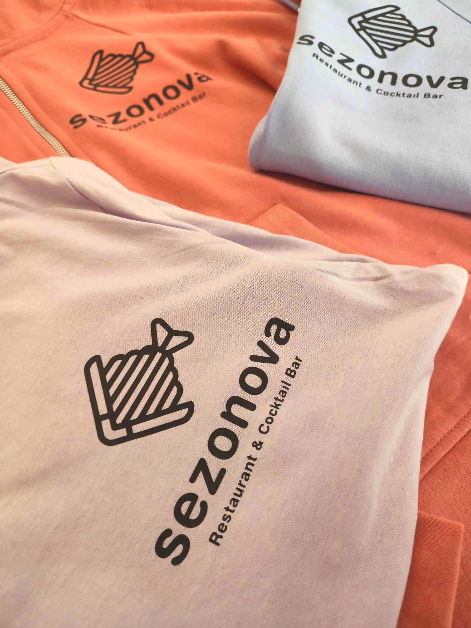 Trzy koszulki w odcieniach pastelowych (róż, fiolet, błękit) z nadrukowanym logo restauracji i baru koktajlowego 'sezonova'. Ułożone warstwowo, widoczne detale nadruku.