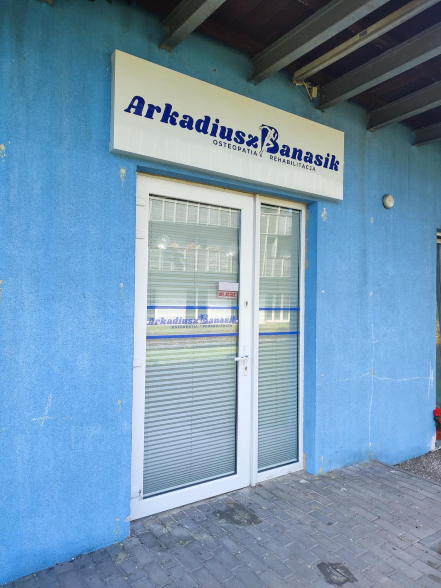 Wejście do gabinetu osteopatii i rehabilitacji Arkadiusz Banasik, z tablicą informacyjną nad drzwiami, na tle niebieskiej elewacji budynku.