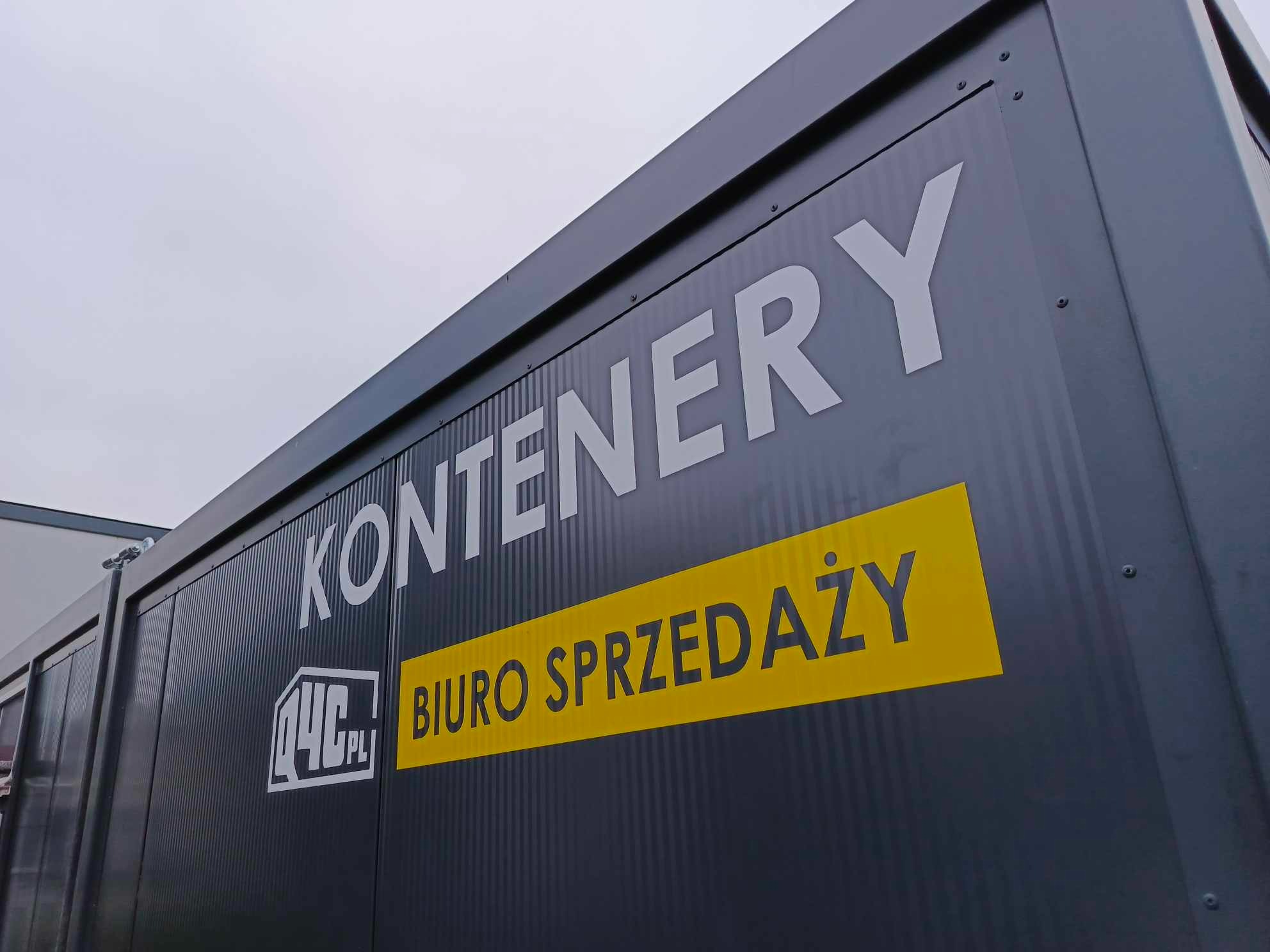 Kontener biurowy z napisem 'Kontenery Biuro Sprzedaży' w kolorach białym i żółtym na szarym tle, widok z boku, z lekkiego dołu, pochmurne niebo.