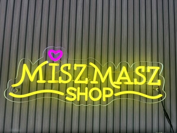 Neonowy szyld 'MISZMASZ SHOP' z różowym sercem, umieszczony na tle pionowych, ciemnych paneli ściennych. Żółte światło neonu oświetla napis.