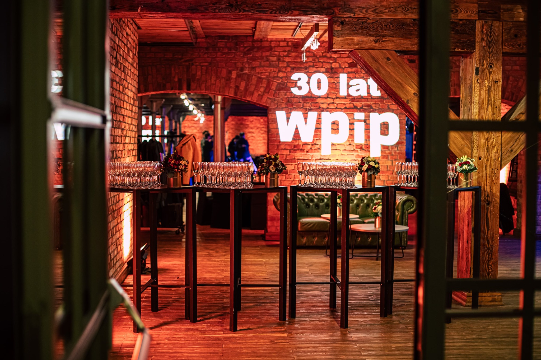 Uroczysta sala z ceglaną ścianą, podświetlonym napisem '30 lat wpip', stolikami z kieliszkami i kwiatami, widziana przez otwarte drzwi.