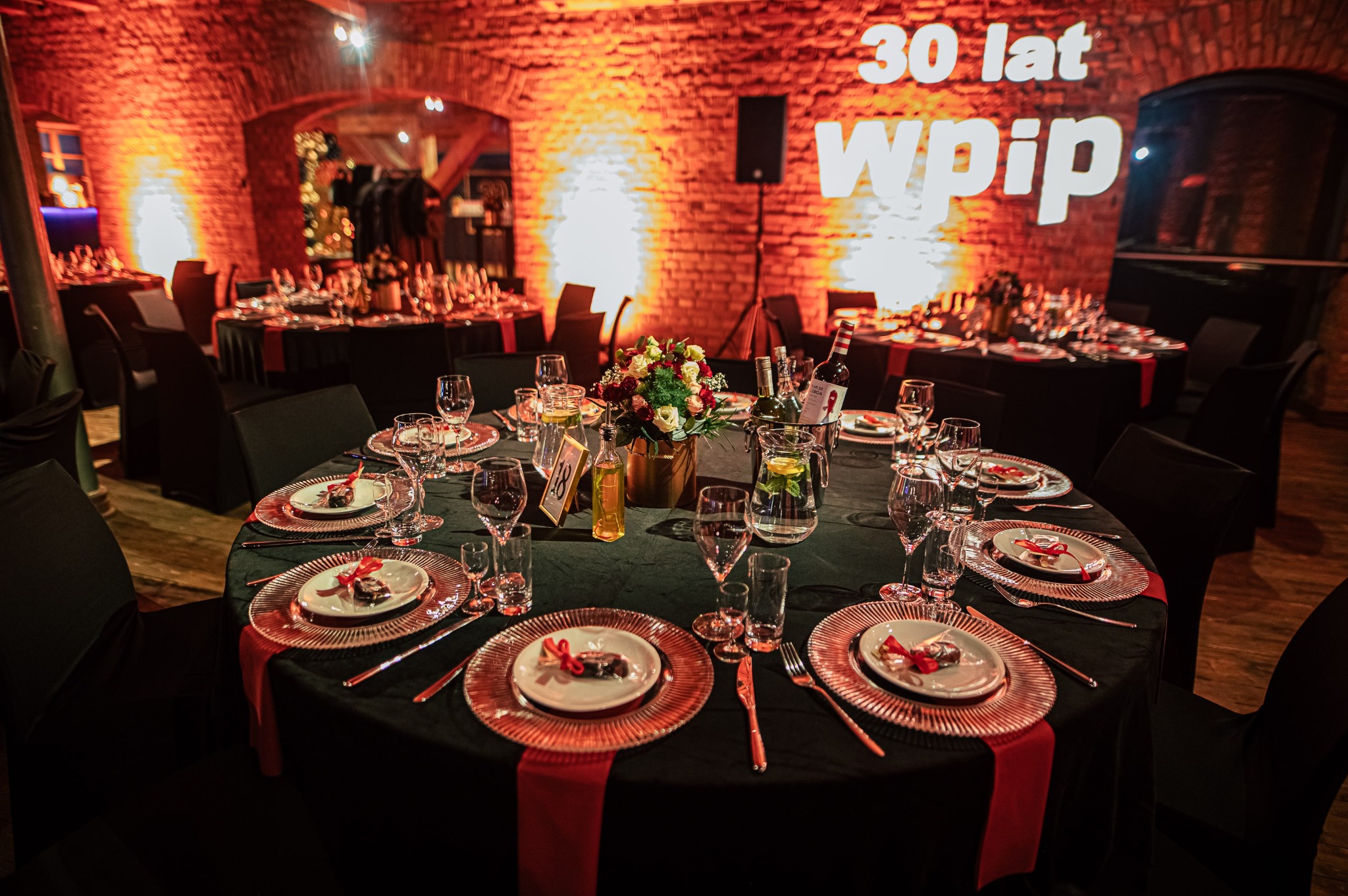 Uroczysta aranżacja stołu w Jasin: czarne obrusy, złote talerze, eleganckie szkło, kompozycje kwiatowe i dekoracje świetlne na ceglanej ścianie.