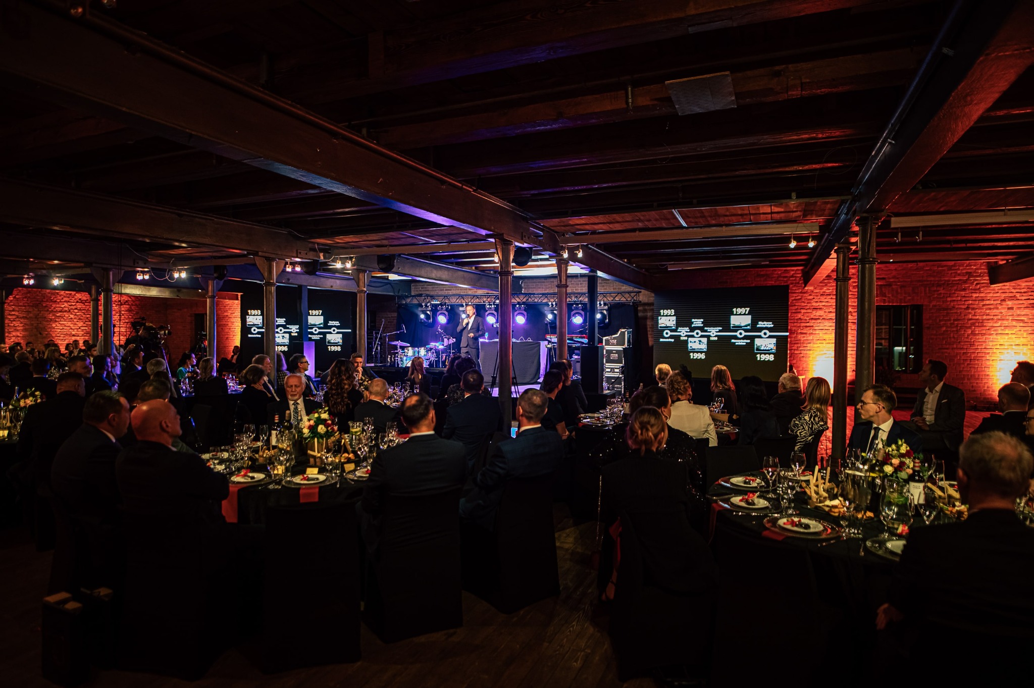 Uroczysta kolacja firmowa w loftowym wnętrzu: eleganckie stoły z kwiatami, scena z występem, ceglane ściany z projekcjami. Oświetlenie punktowe tworzy intymną atmosferę.