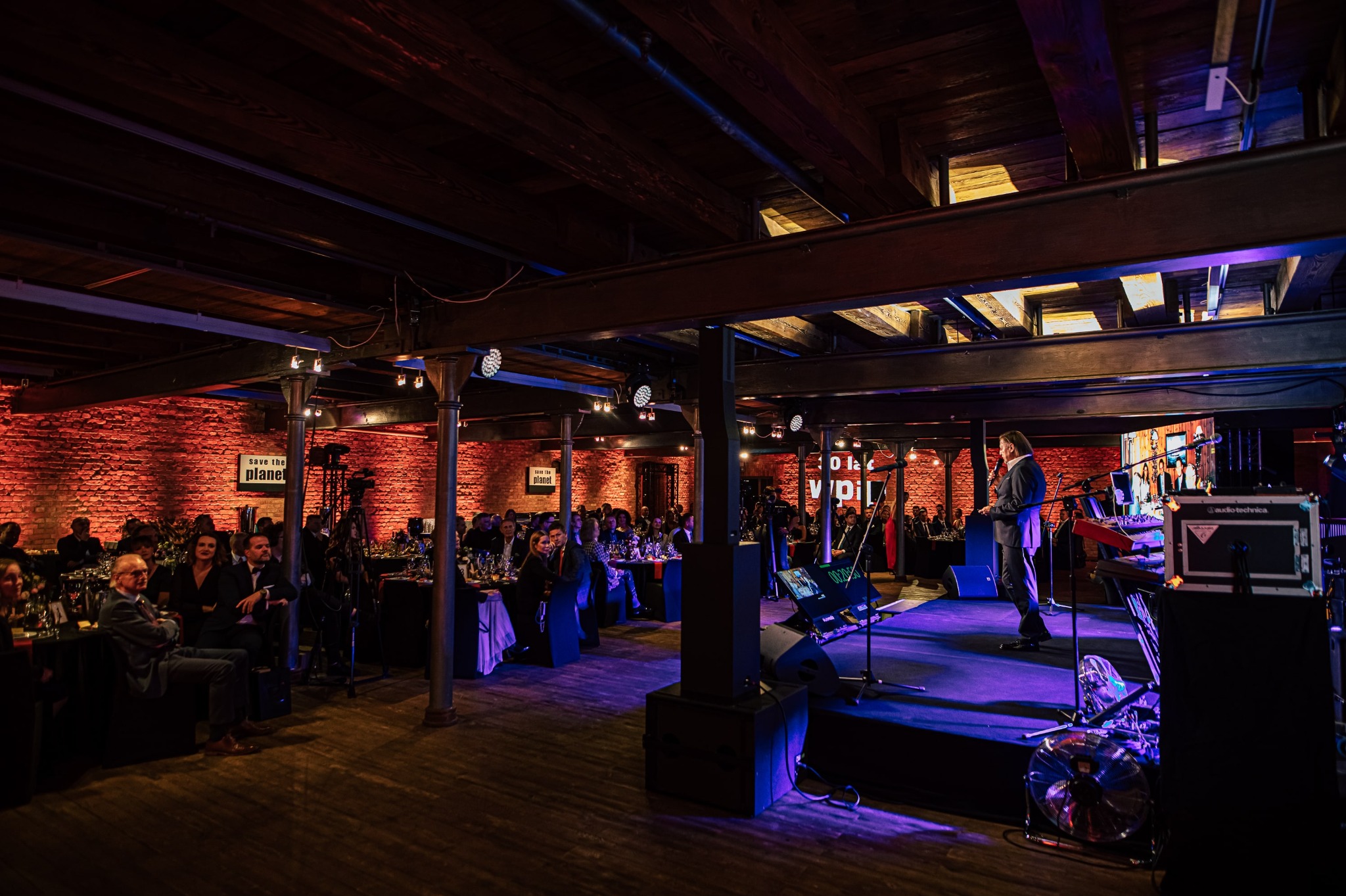 Sala eventowa z ceglanymi ścianami, oświetlona na czerwono i niebiesko. Goście siedzą przy stołach, a na scenie występuje prelegent. Widoczny sprzęt nagłośnieniowy.