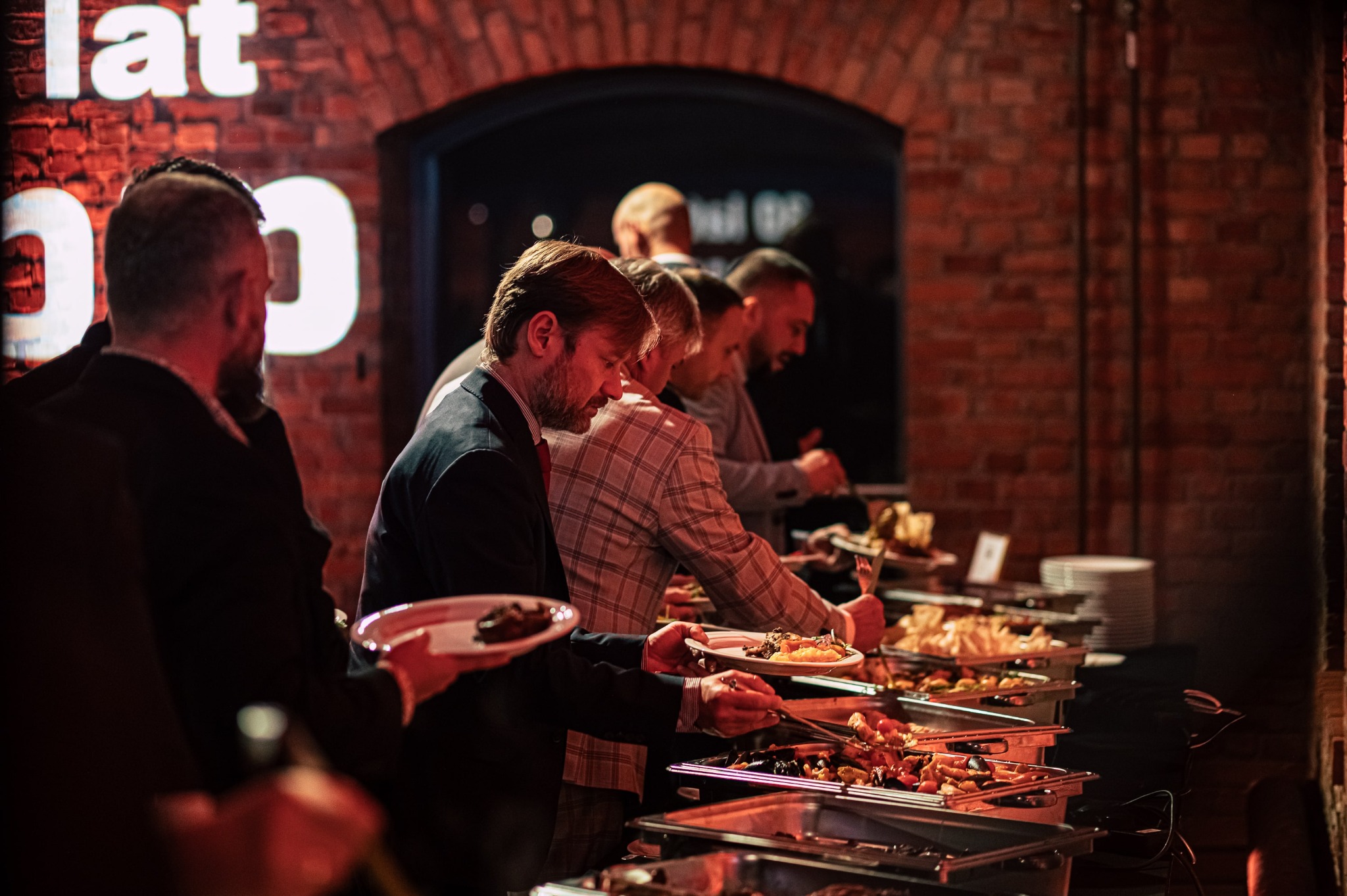 Elegancki bufet z różnorodnymi daniami podczas eventu firmowego. Goście w garniturach nakładają jedzenie na talerze, ceglane ściany i ciepłe oświetlenie tworzą wyjątkową atmosferę.