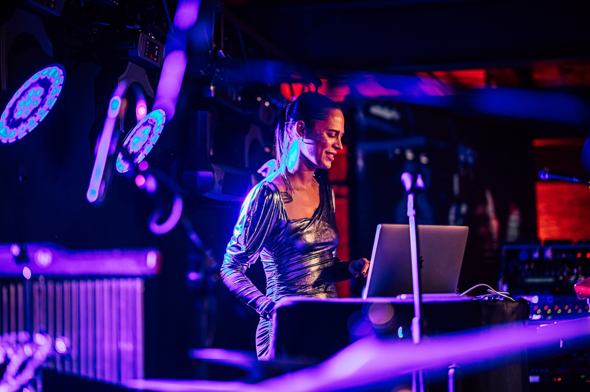 Kobieta DJ w srebrnej sukience, grająca muzykę na laptopie podczas eventu, oświetlona fioletowymi światłami scenicznymi z efektem bokeh.