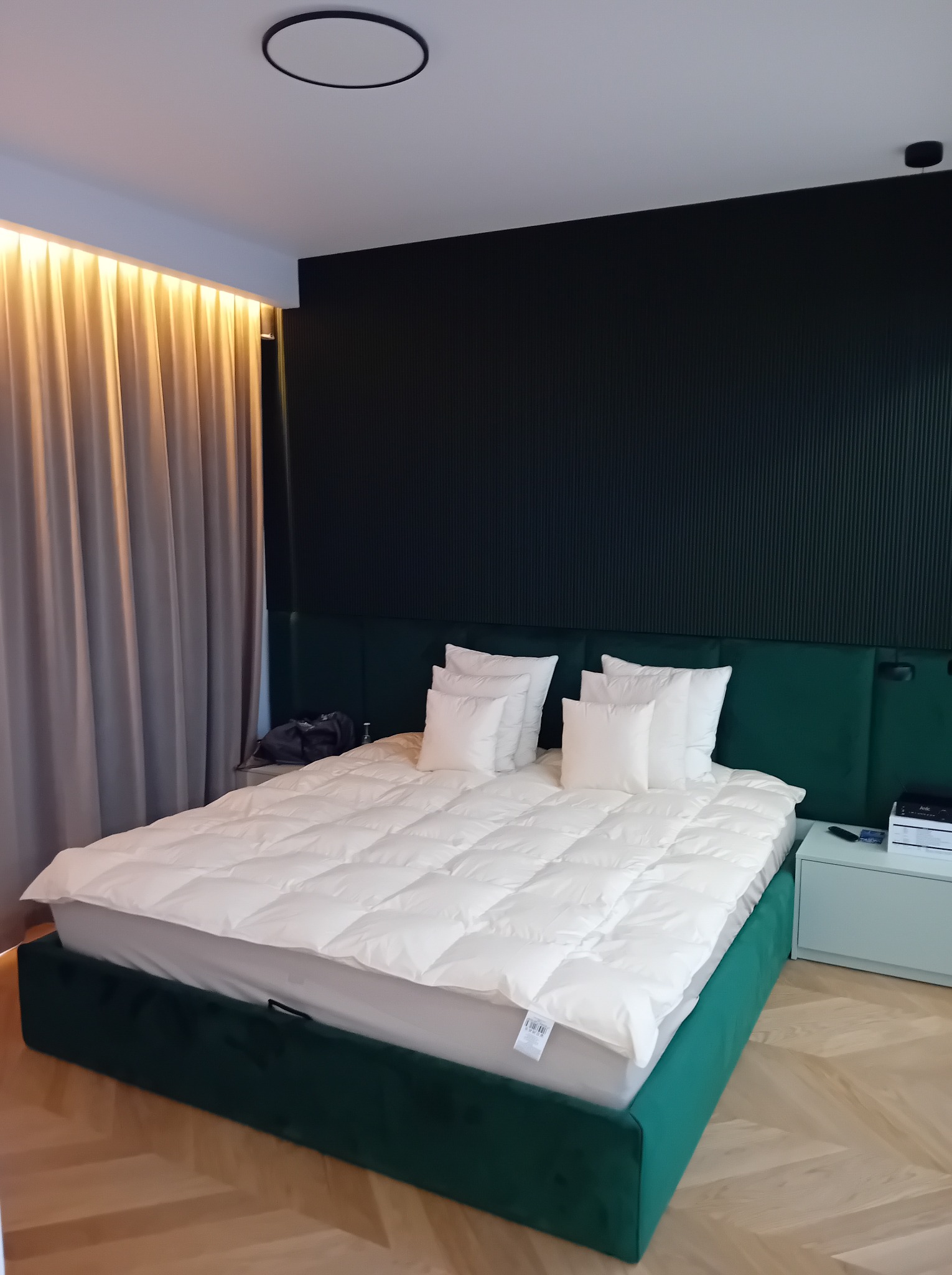 Nowoczesna sypialnia z zielonym łóżkiem i zagłówkiem, białą pościelą, ciemną ścianą z pionowymi panelami i drewnianą podłogą w jodełkę. Minimalistyczny design.