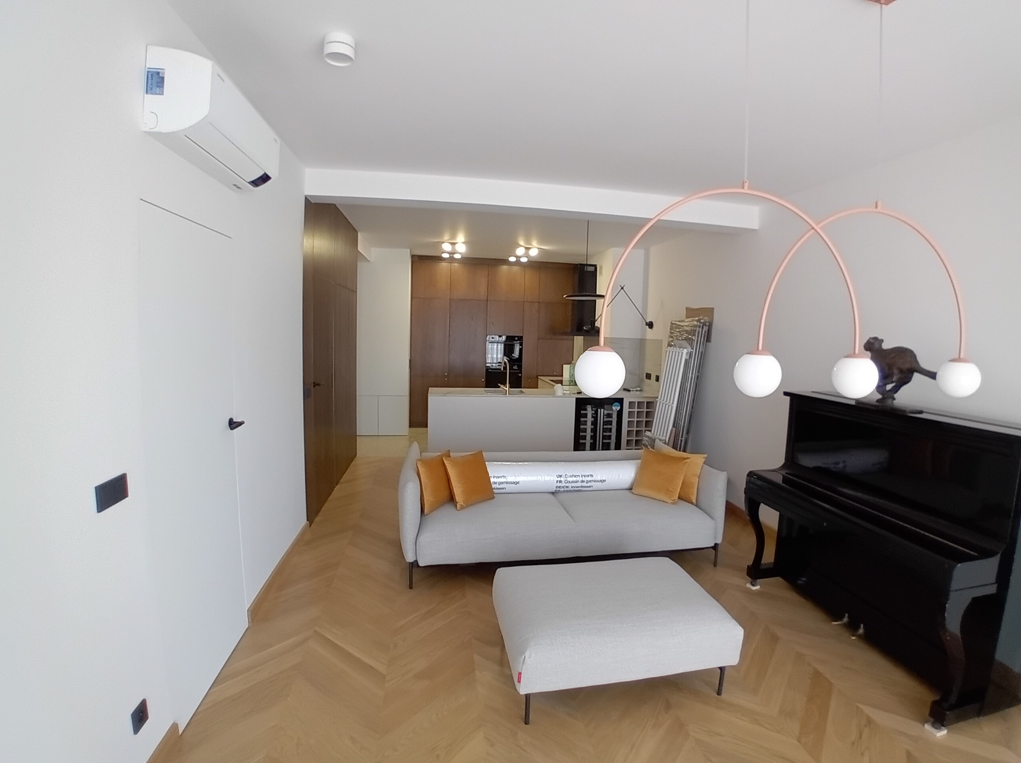 Salon z parkietem w jodełkę, szarą sofą z pomarańczowymi poduszkami, fortepianem i nowoczesną lampą. Widok na otwartą kuchnię w tle. Klimatyzator na ścianie.