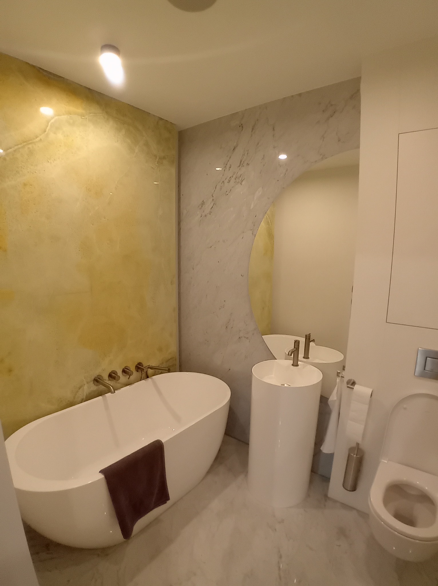 Nowoczesna łazienka z wolnostojącą wanną, umywalką na postumencie i toaletą. Ściany wykończone marmurem i imitacją onyksu. Minimalistyczny design, ciepłe oświetlenie.