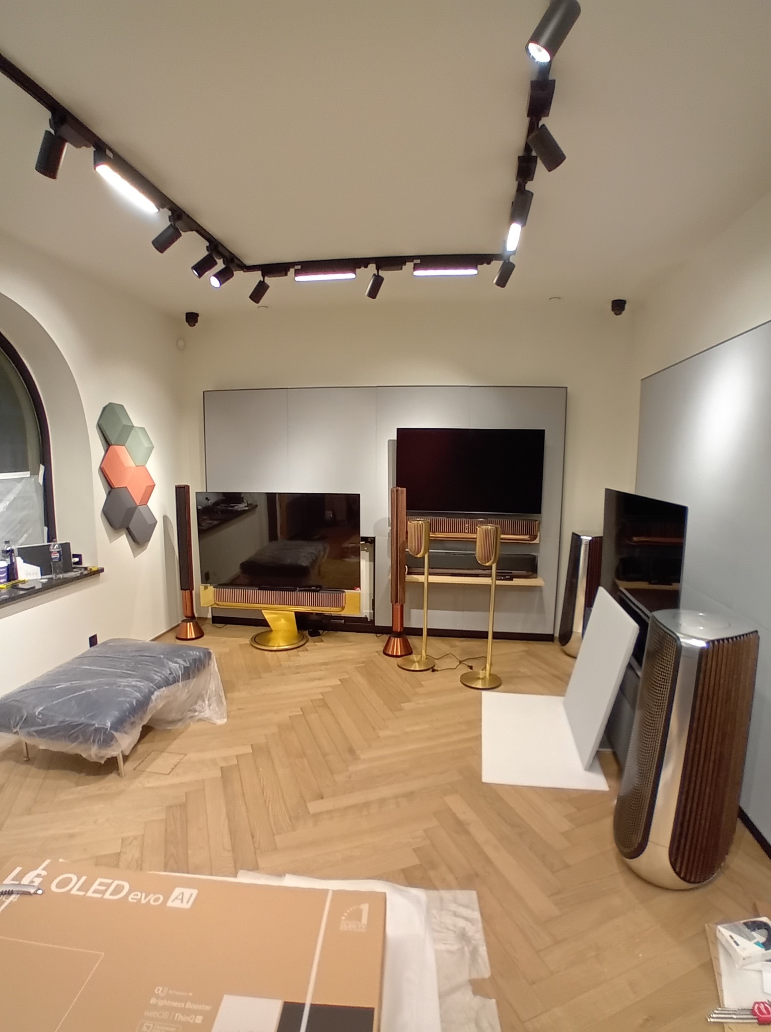 Salon z parkietem w jodełkę, oświetleniem szynowym i elementami audio-video Bang & Olufsen. Część mebli zabezpieczona folią. W tle heksagonalne panele ścienne.