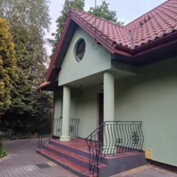 Elewacja domu w pastelowej zieleni z kolumnami, czerwonym dachem i ozdobnymi barierkami. Widoczne schody wejściowe i fragment ogrodu z drzewami.