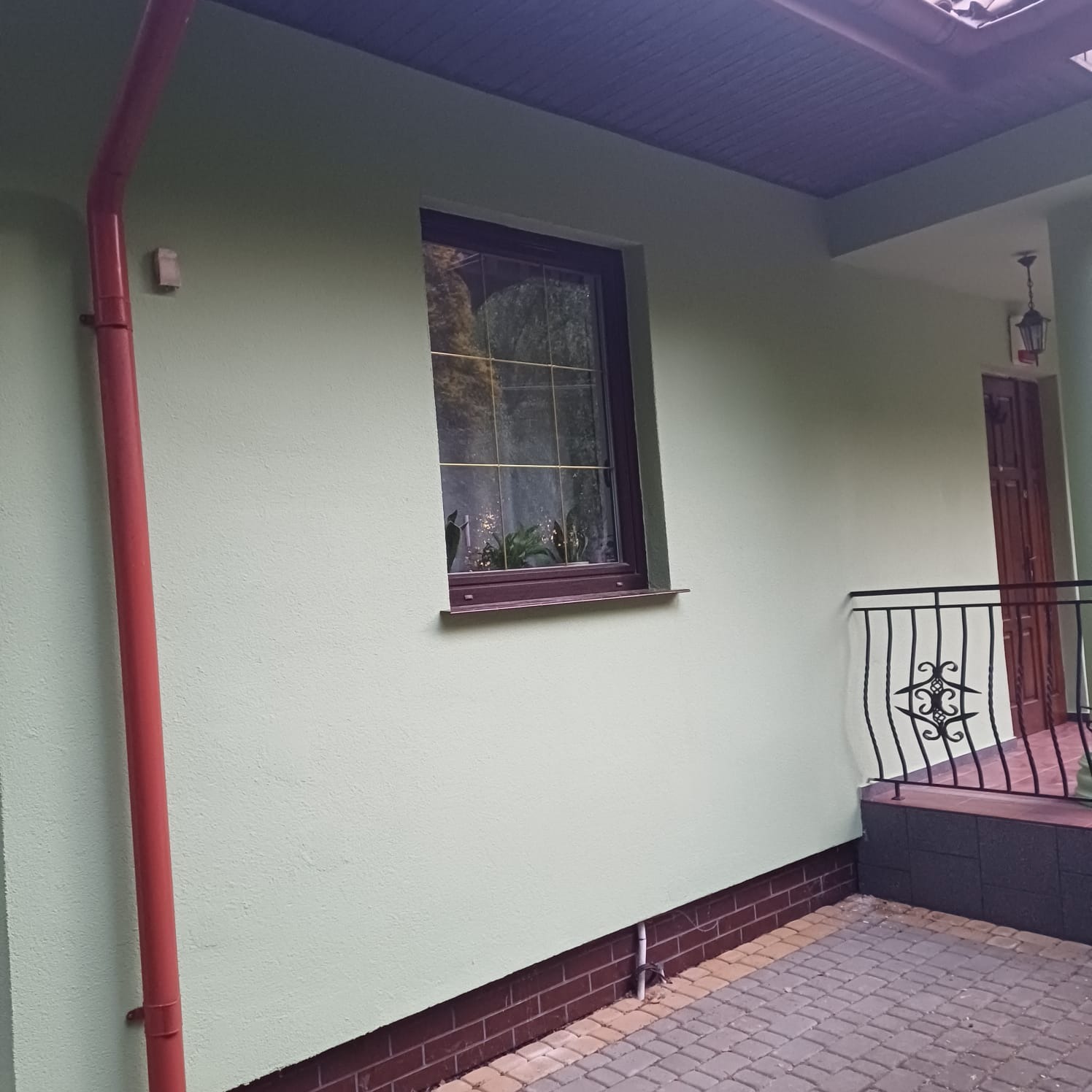 Ściana domu po malowaniu na jasnozielony kolor, z brązowymi detalami: okno, cokół i rynna w kolorze czerwonym. Widoczny fragment kostki brukowej.