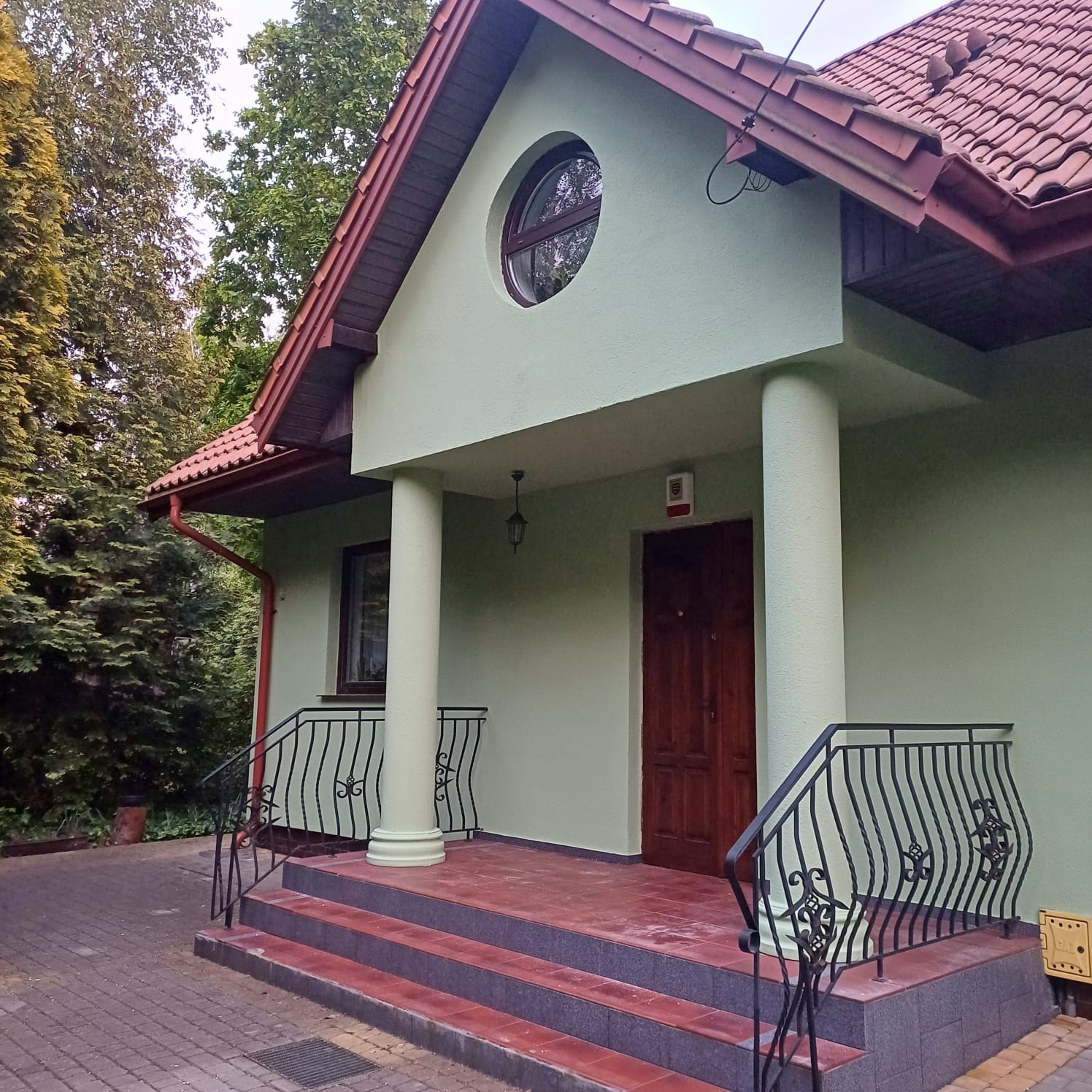 Elewacja domu jednorodzinnego po renowacji: pastelowa zieleń ścian, ciemna stolarka okienna i drzwi, czerwona dachówka, okrągłe okno na poddaszu, ozdobne balustrady.