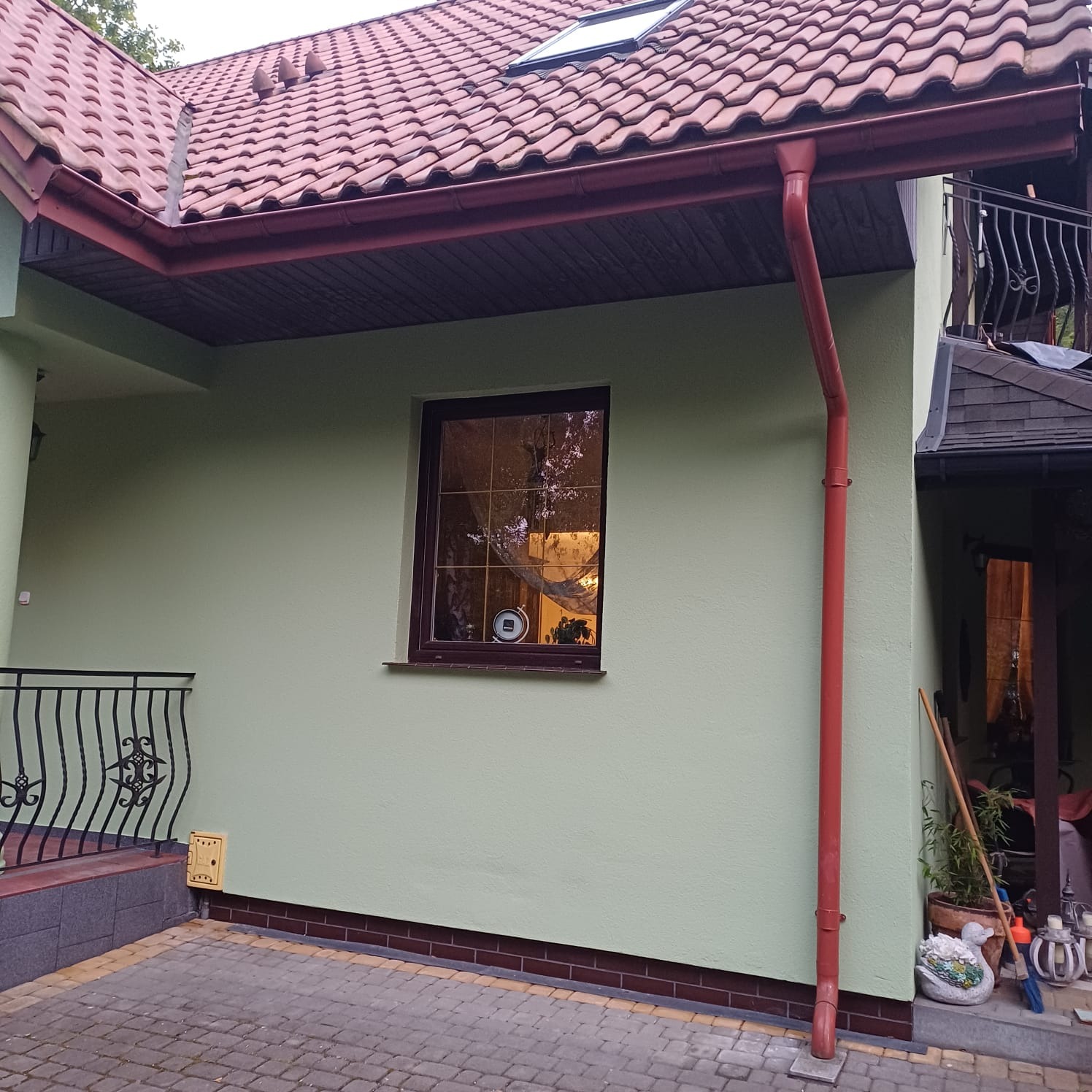 Jasnozielona elewacja domu z brązową stolarką okienną i rynną, widoczny fragment dachu z dachówką i kostka brukowa na podjeździe.