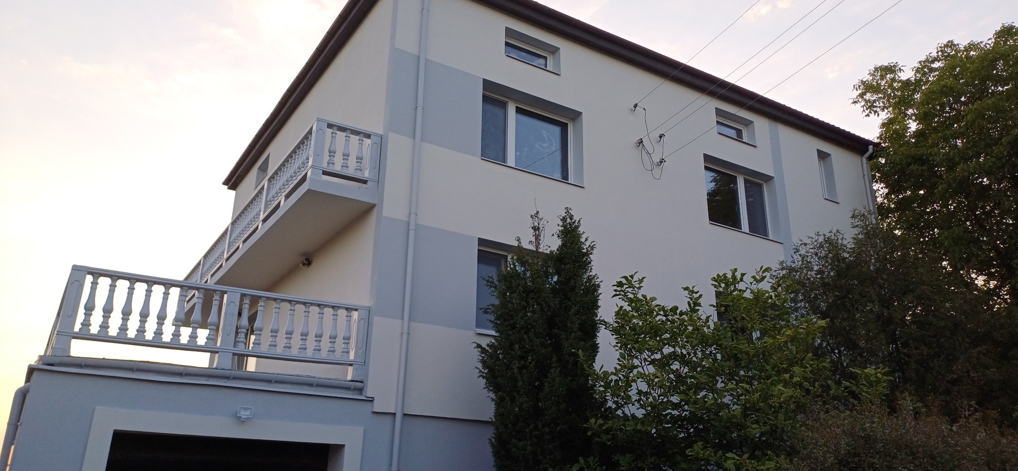 Dwukondygnacyjny dom po odświeżeniu elewacji w odcieniach szarości i bieli, z balkonem z balustradą i widocznymi przewodami elektrycznymi na ścianie. Zieleń drzew w tle.