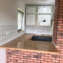 Kuchenny blat drewniany z oświetleniem i ceglaną ścianą, widok na białe szafki. Świeże wykończenie, płytki na ścianie z gniazdkami, surowe drewno na suficie.