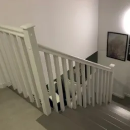 Klatka schodowa z szarymi schodami i białą balustradą. Na ścianie wiszą obrazy w czarnych ramkach. Widok z góry na dół, oświetlenie górne.