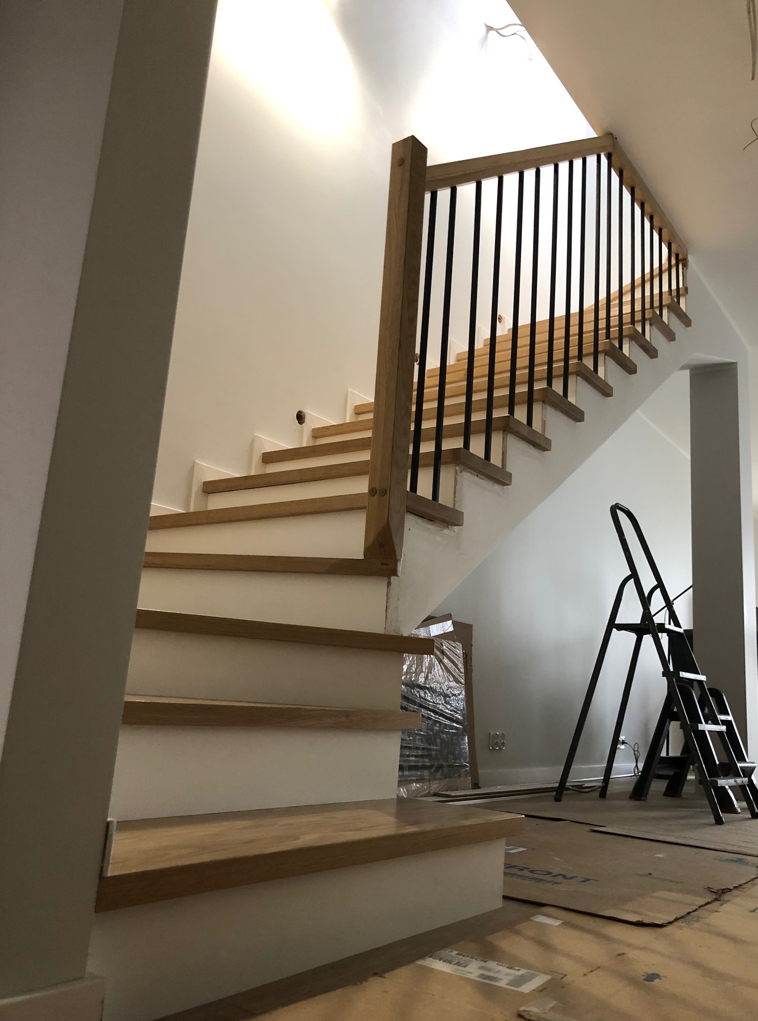 Drewniane schody z czarną balustradą w jasnym wnętrzu. Widoczne zabezpieczenia podłogi i drabina, sugerujące etap wykończeniowy. Naturalne oświetlenie.