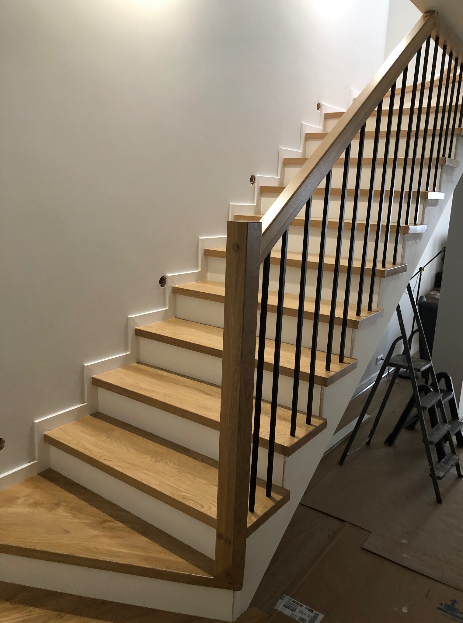 Nowoczesne schody drewniane z czarną balustradą i jasnym drewnem, widok z dołu, z metalową drabiną w tle. Detal wykończenia i naturalne światło.