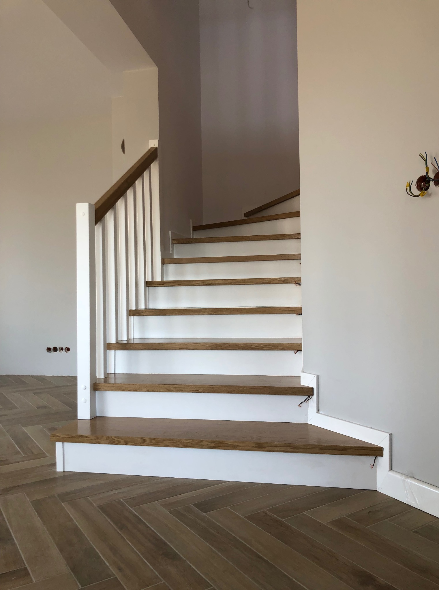 Nowoczesne schody drewniane z białymi podstopnicami i drewnianymi stopniami, jasne wnętrze z parkietem ułożonym w jodełkę, widoczne niedokończone instalacje.