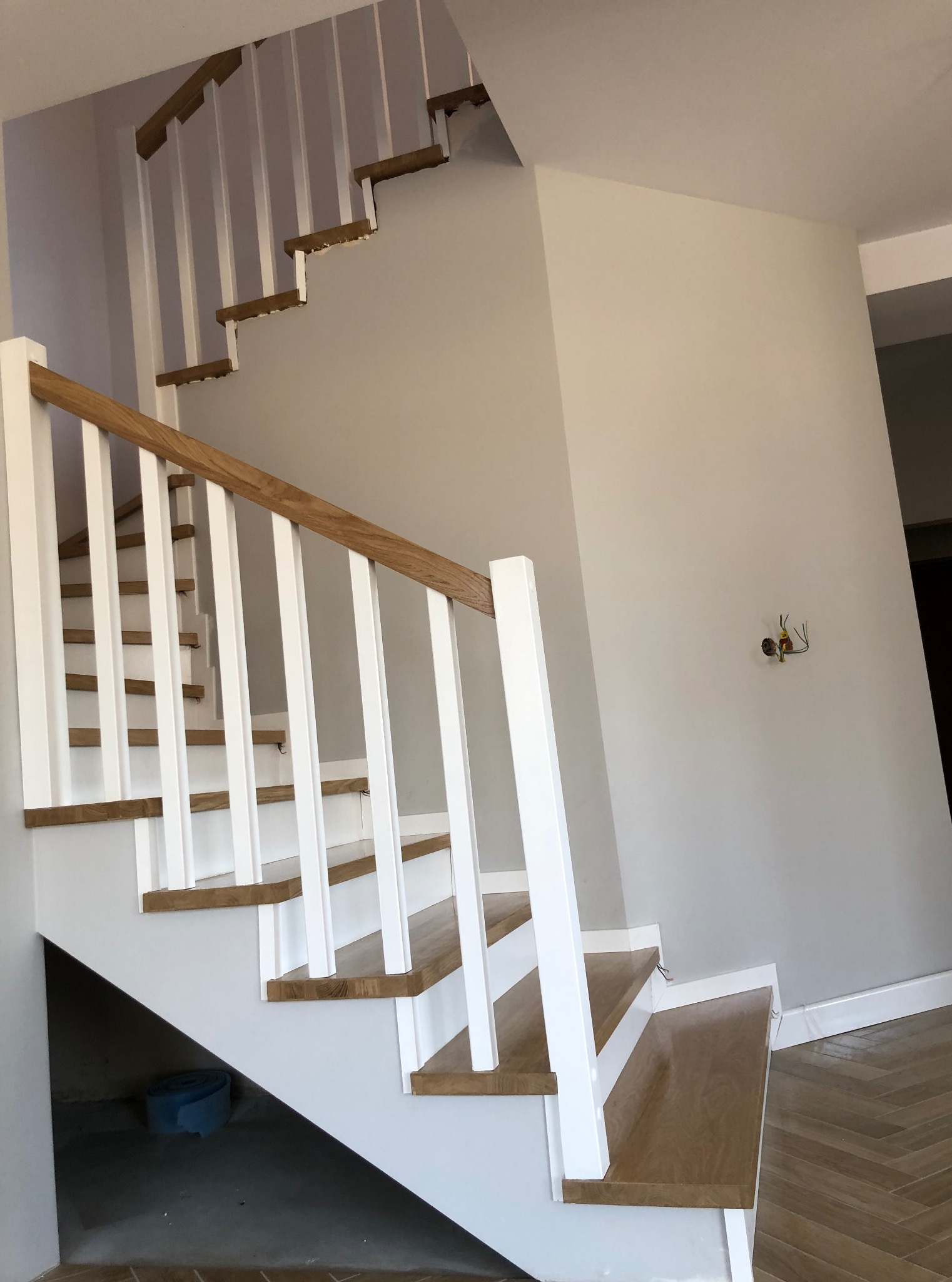 Nowoczesne schody drewniane z białymi balustradami i dębowymi stopniami, widoczne z dołu, z fragmentem podłogi w jodełkę i wystającymi kablami ze ściany.