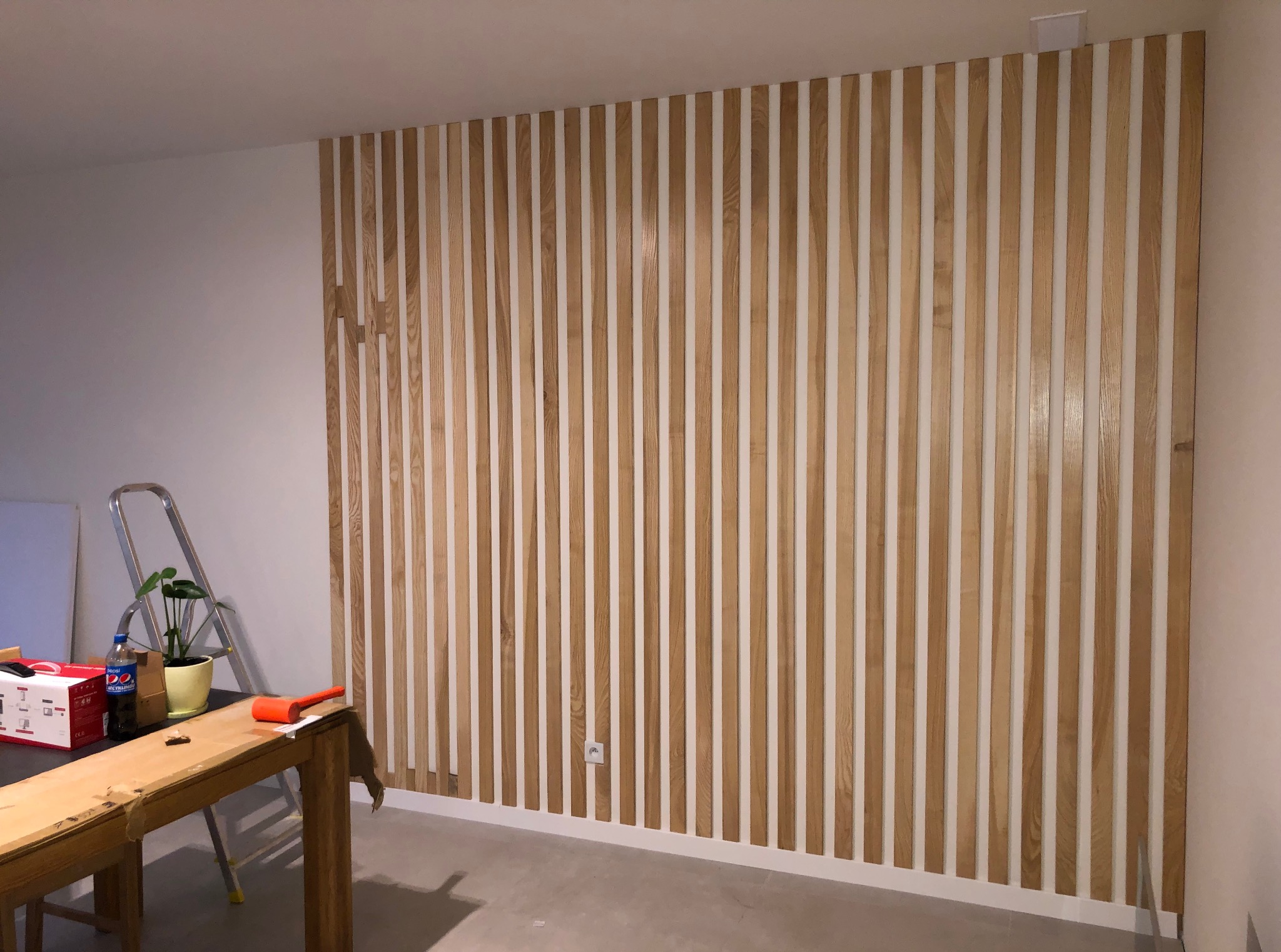 Drewniana dekoracja ścienna z listew w jasnym odcieniu drewna, pionowy układ, białe tło, widoczny fragment pomieszczenia w trakcie wykańczania.