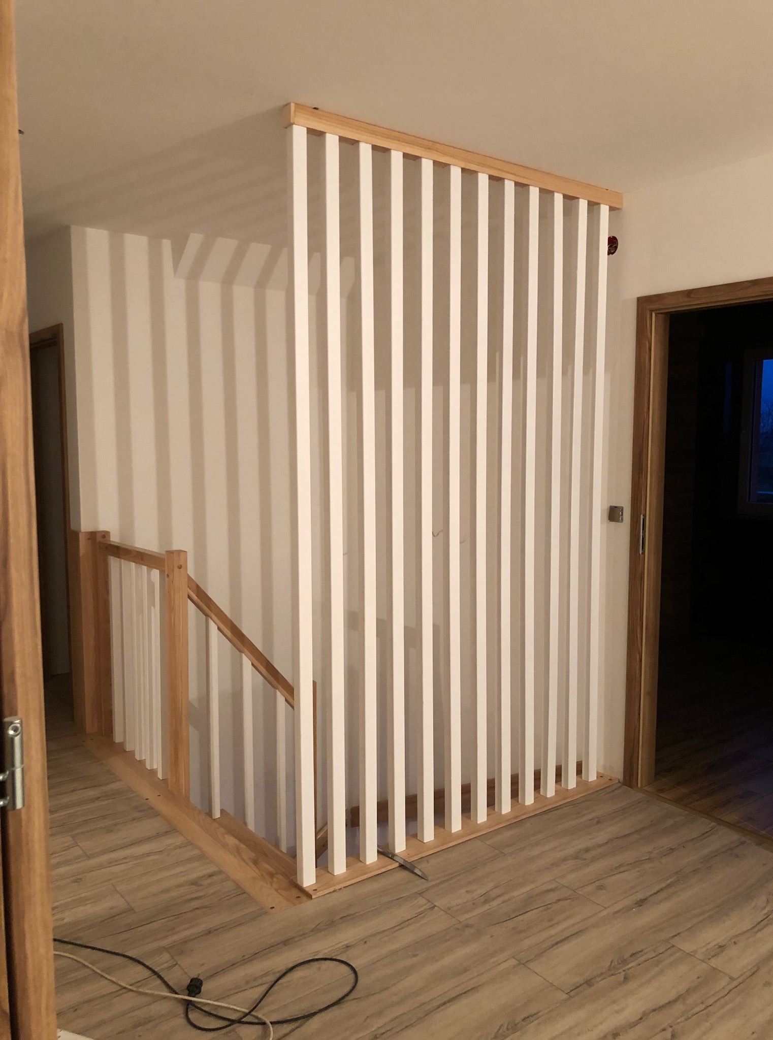 Nowoczesna, ażurowa balustrada schodowa z białymi pionowymi elementami i drewnianą ramą, widok z dołu na jasne wnętrze z drewnianą podłogą.