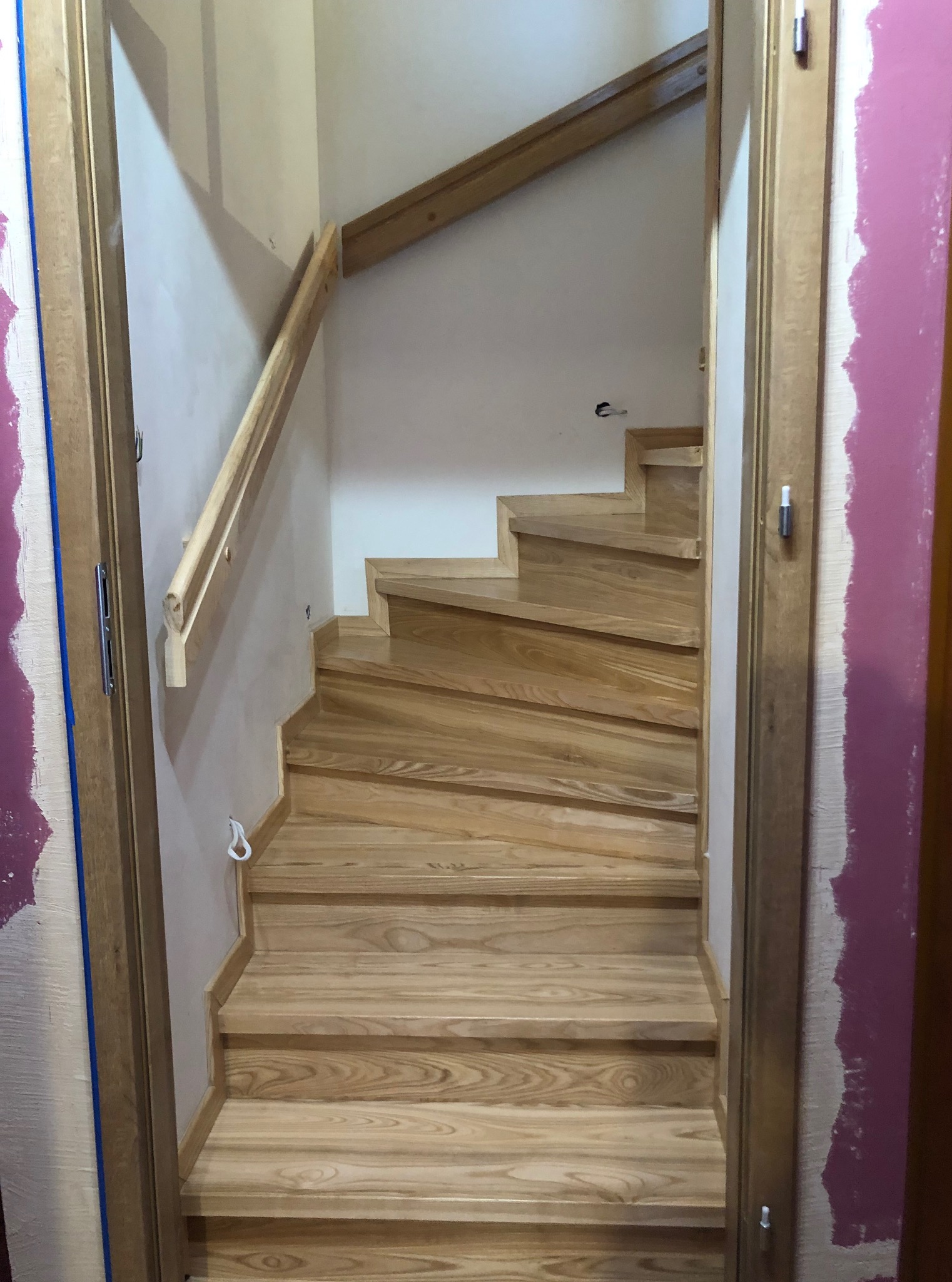 Jasne, drewniane schody z poręczą, widziane z dołu, podczas montażu. Ściany w trakcie malowania. Elementy wykończeniowe w kolorze drewna.