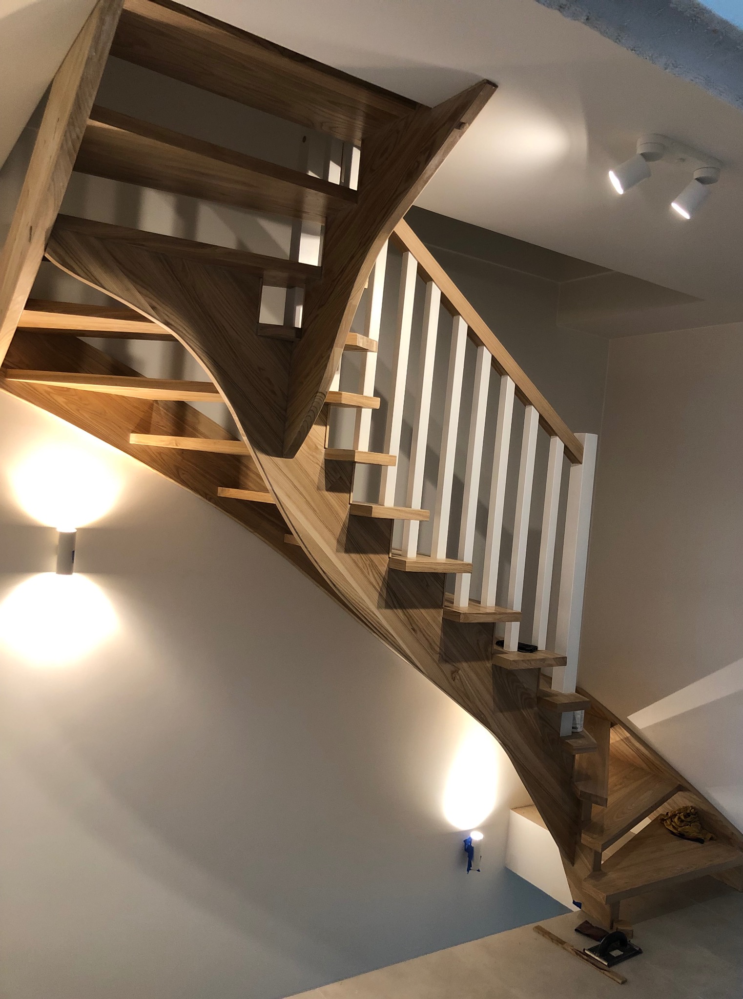 Nowoczesne schody z drewna z białą balustradą, oświetlone punktowo, z widocznym zakrzywionym biegiem i detalami wykończenia. Jasne drewno, minimalistyczny design.