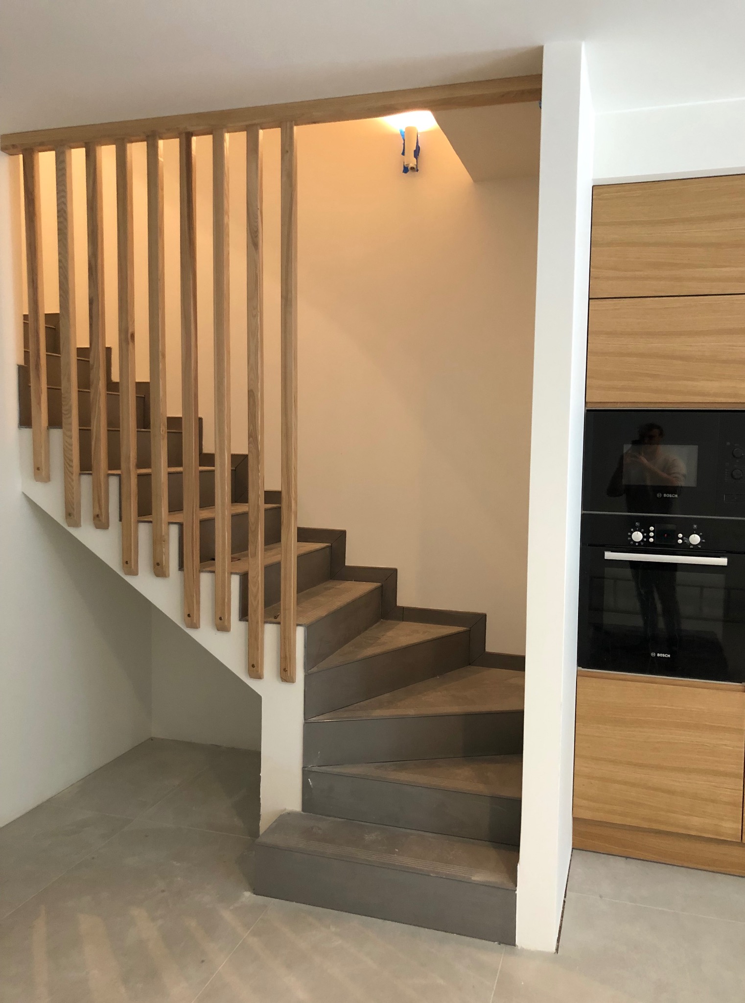 Nowoczesne schody z drewnianą balustradą w pionowe listwy, szare stopnie, zabudowa kuchenna z drewnianymi frontami w tle. Widok z dołu, oświetlenie sufitowe.