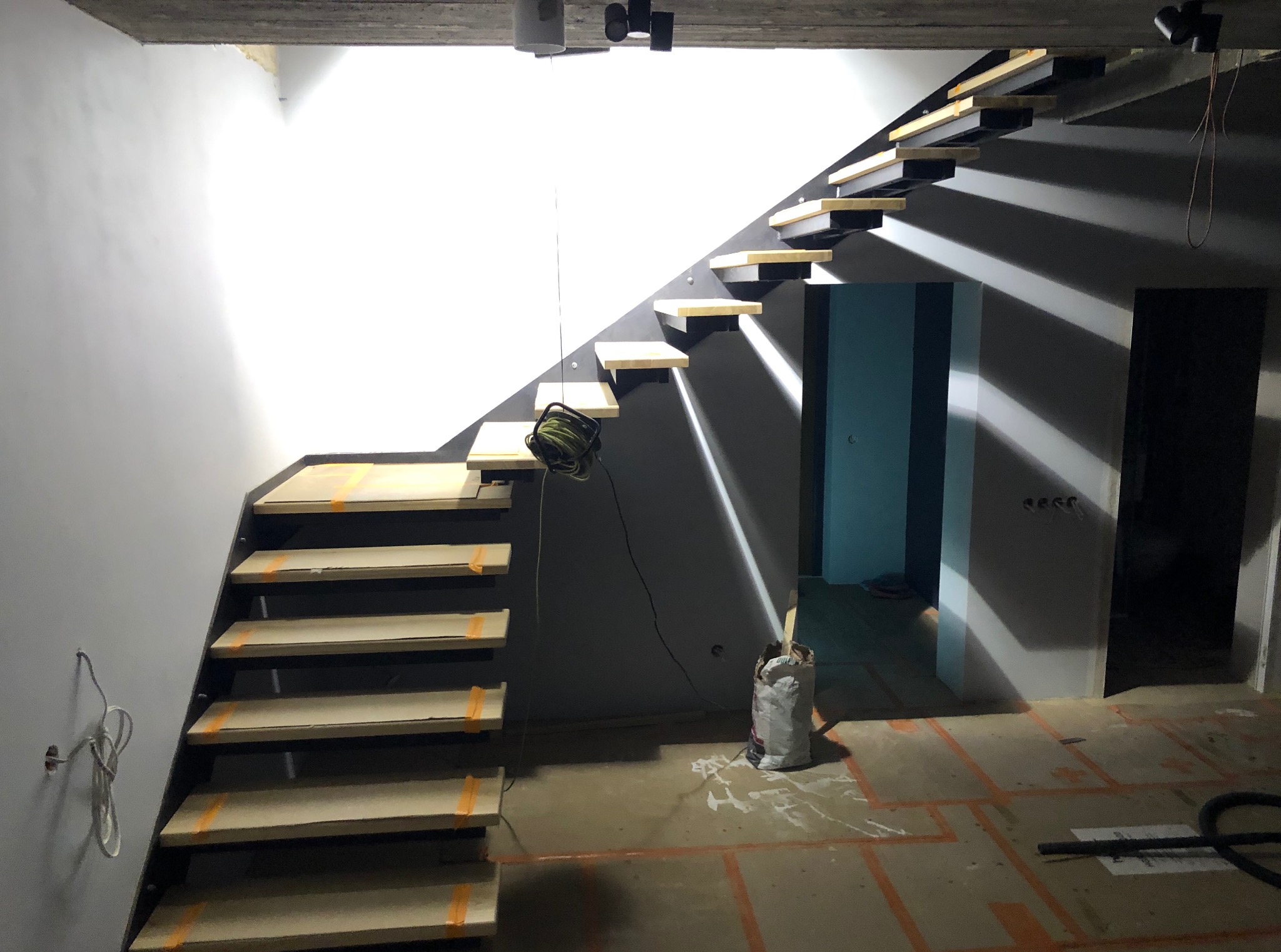 Nowoczesne schody z drewnianymi stopniami w surowym wnętrzu. Stopnie zabezpieczone taśmą, widoczny kabel i worek, co sugeruje etap wykończenia.