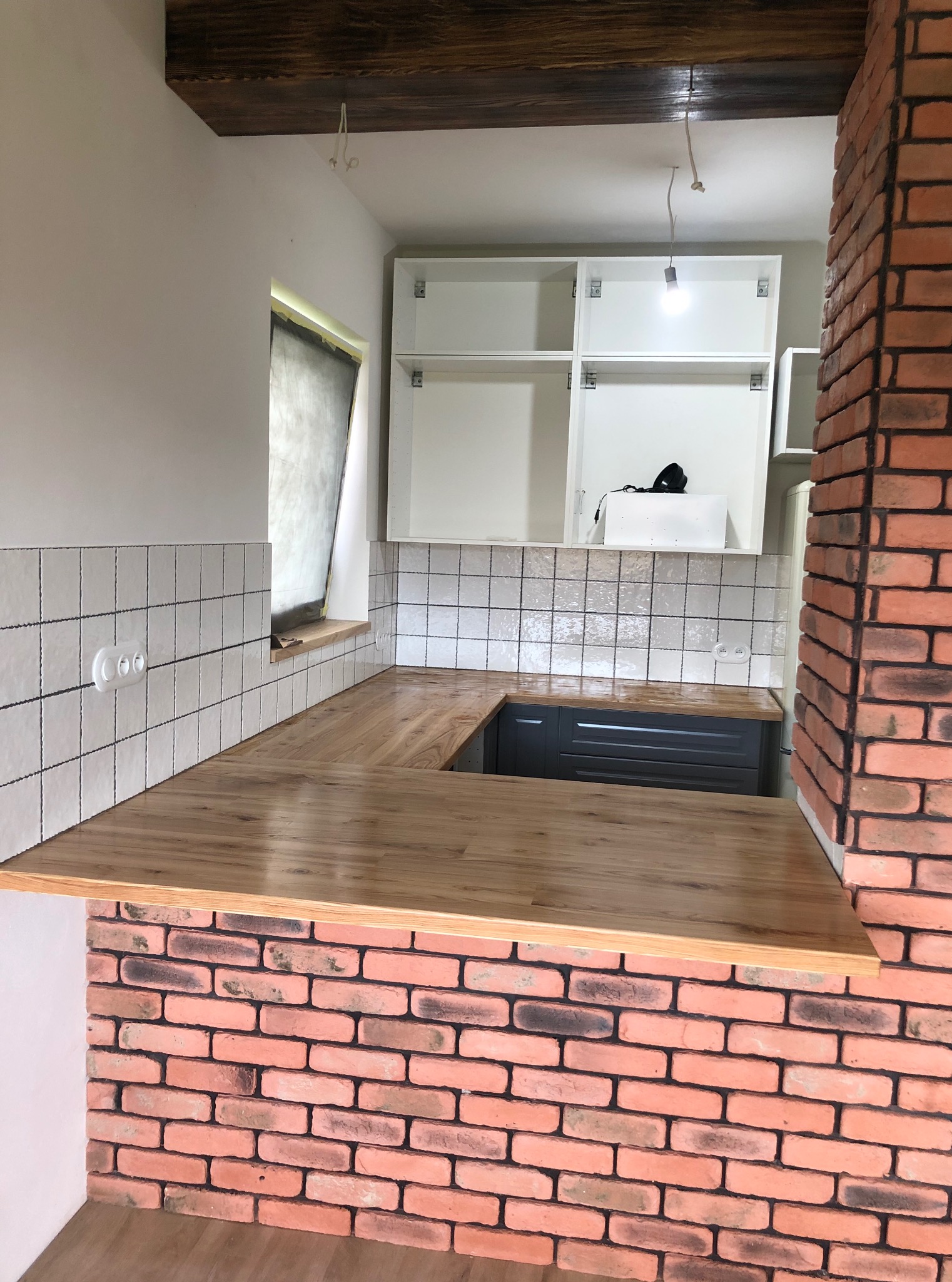 Kuchenny blat drewniany z oświetleniem i ceglaną ścianą, widok na białe szafki. Świeże wykończenie, płytki na ścianie z gniazdkami, surowe drewno na suficie.