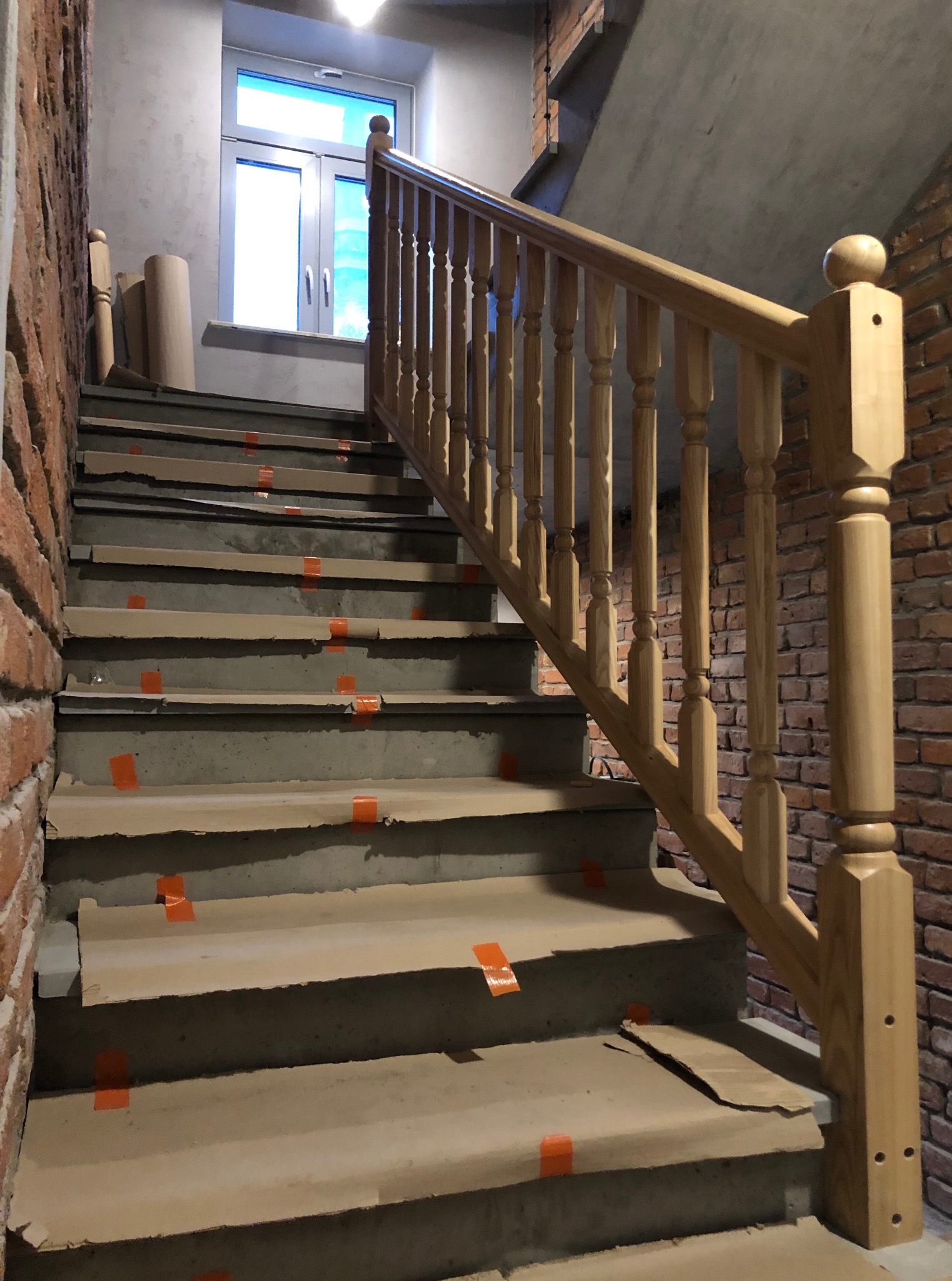 Surowe betonowe schody zabezpieczone kartonem i taśmą, z jasną, drewnianą balustradą z okrągłym zakończeniem, na tle ściany z cegły i okna.