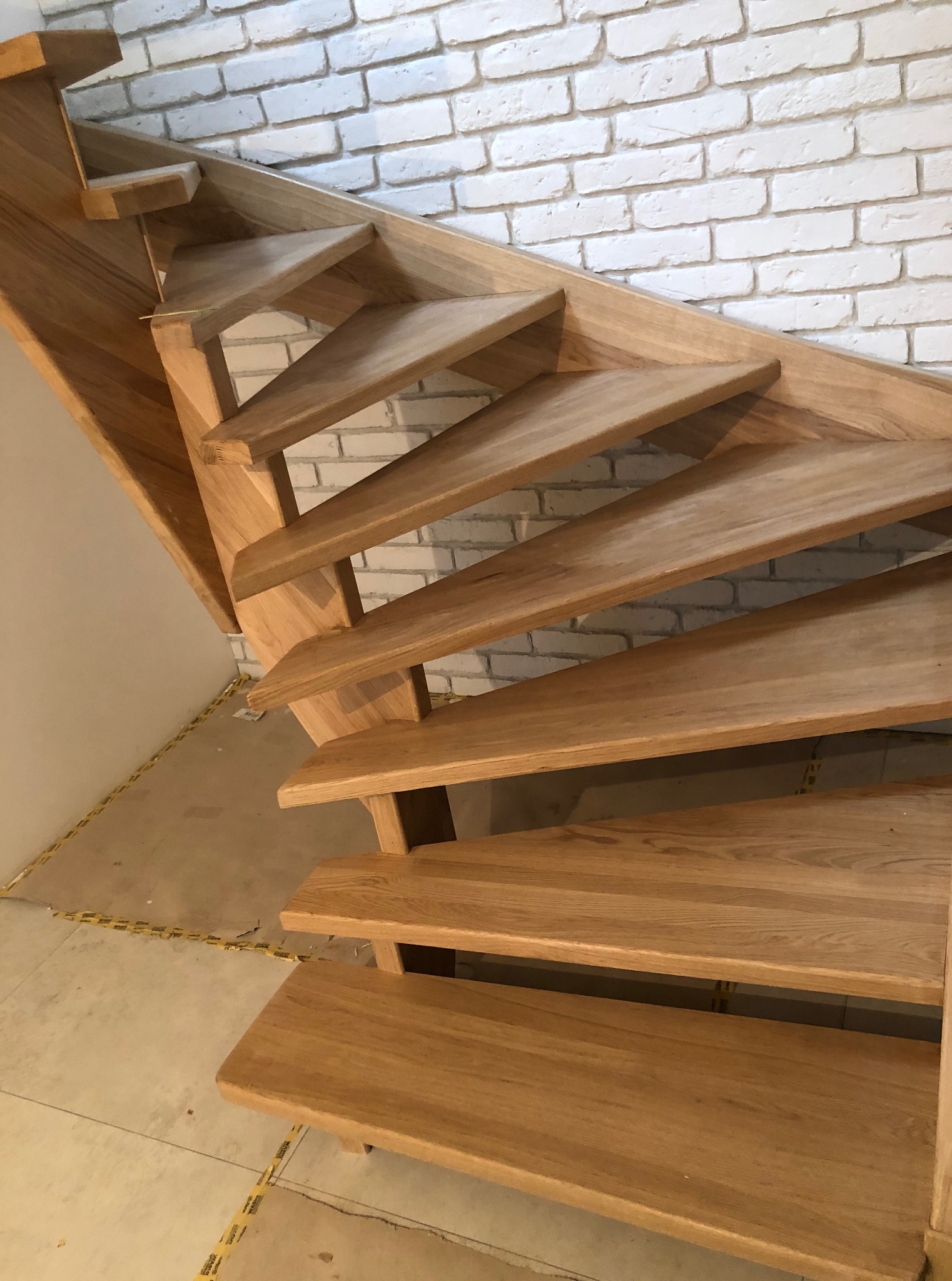 Drewniane schody o naturalnym odcieniu, z widoczną strukturą drewna, na tle białej ściany z cegły. Ujęcie z dołu, eksponujące stopnie i konstrukcję.