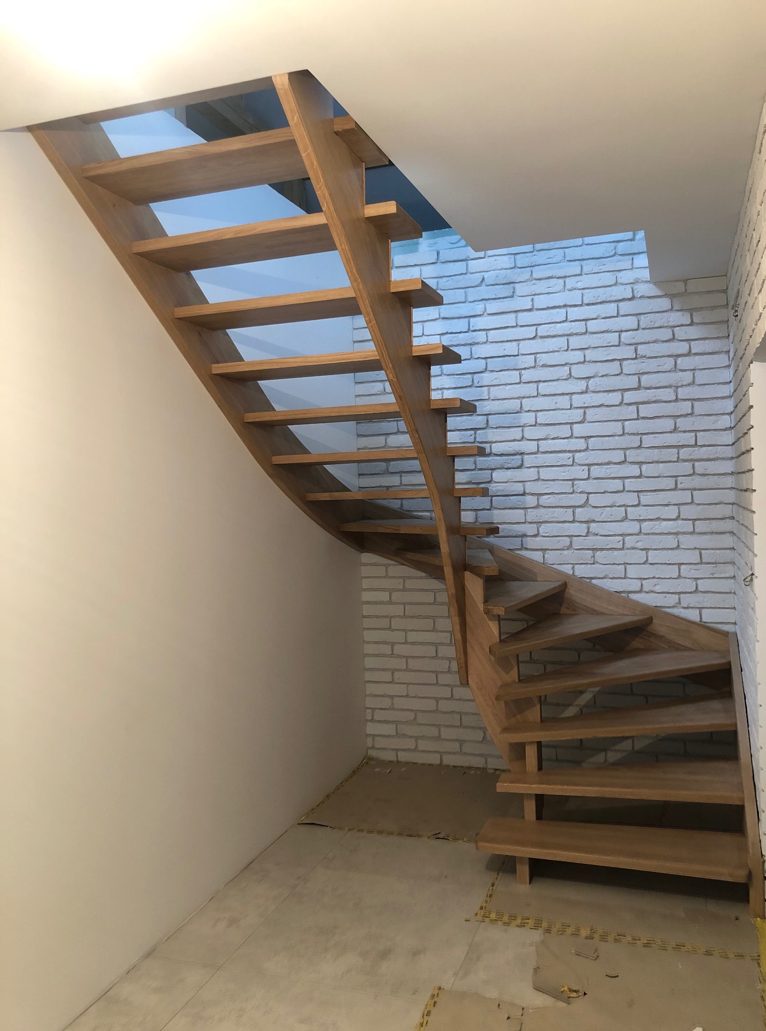 Spiralne schody z jasnego drewna, z widocznymi stopniami i konstrukcją, na tle białej ściany z cegły. Ujęcie z dołu, podkreślające zakrzywiony kształt.