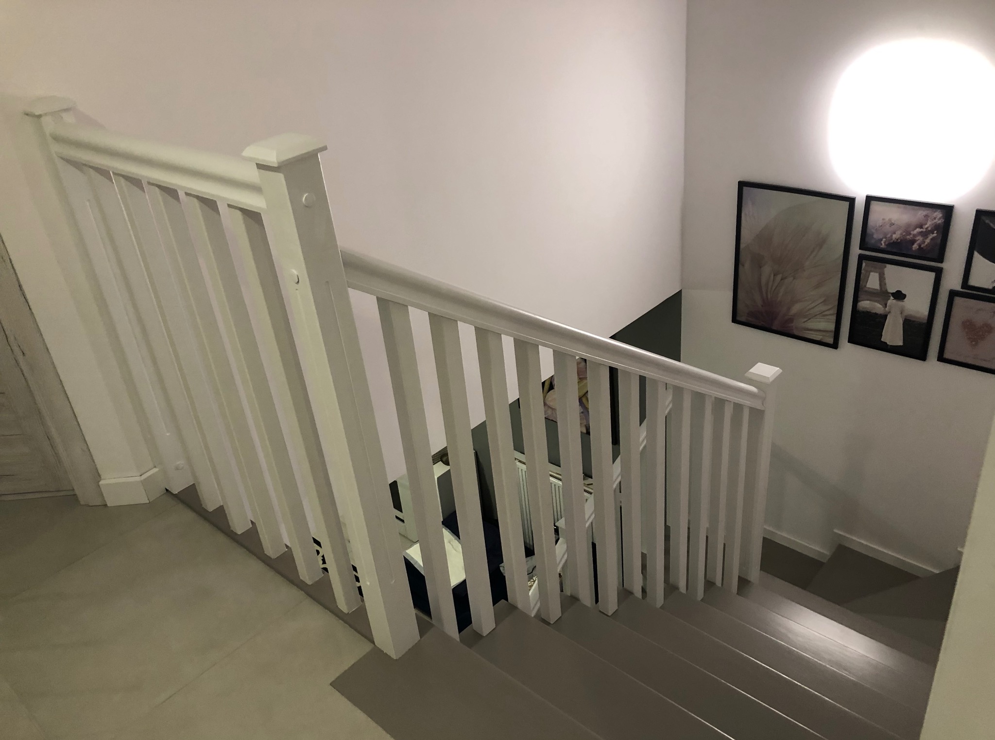 Klatka schodowa z szarymi schodami i białą balustradą. Na ścianie wiszą obrazy w czarnych ramkach. Widok z góry na dół, oświetlenie górne.