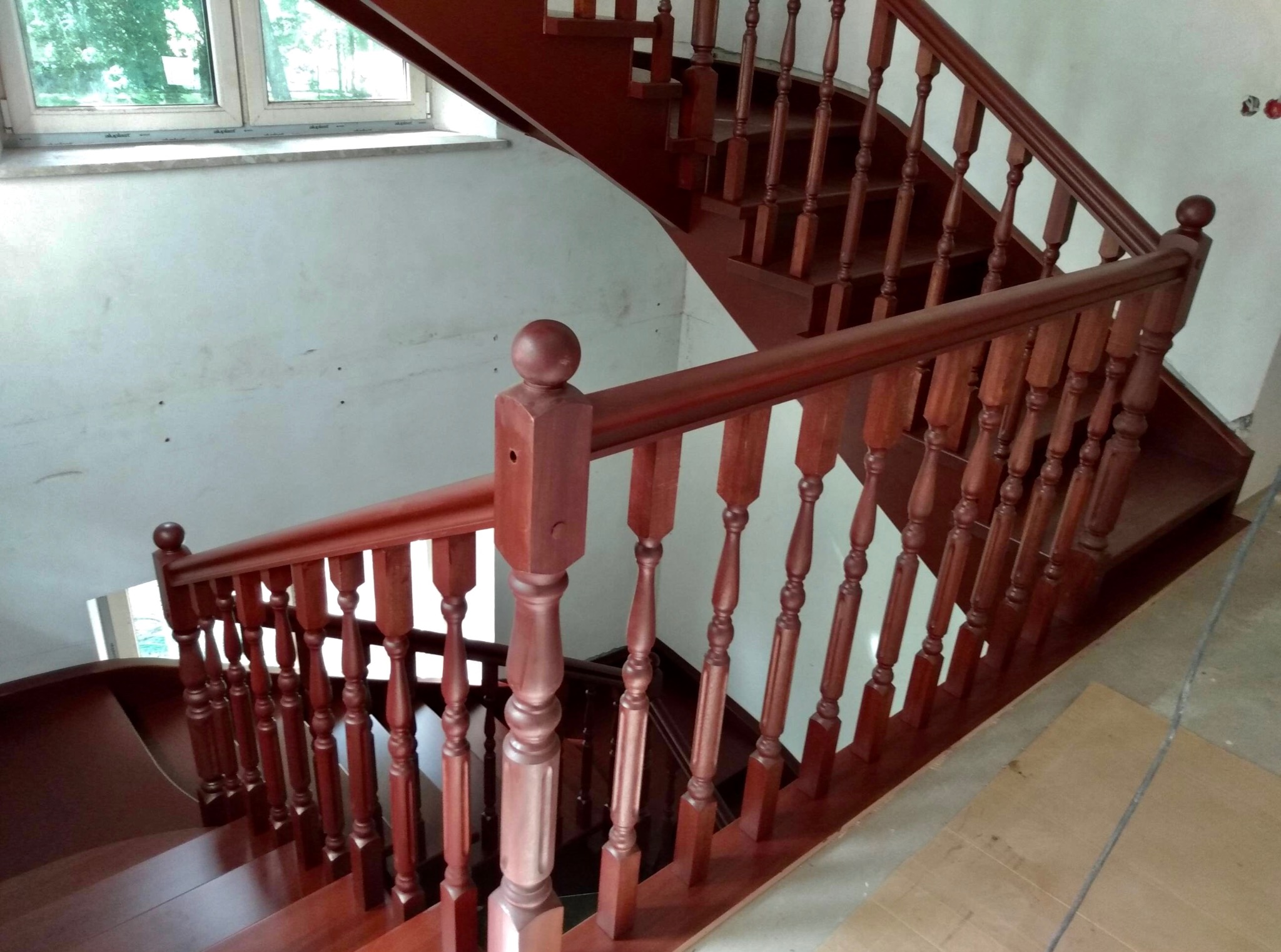 Wewnętrzne, drewniane schody w odcieniach czerwieni, z balustradą z toczonymi elementami. Widok z góry, częściowo na tle okna. Nie wykończone ściany.