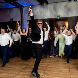 OLSZEWSKI - DJ i Konferansjer - Rozbawiony wokalista w okularach przeciwsłonecznych, z mikrofonem w dłoni, w otoczeniu tańczącego tłumu na parkiecie. Uczestnicy w strojach wieczorowych.