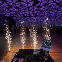 OLSZEWSKI - DJ i Konferansjer - Para młoda tańczy na parkiecie, otoczona fontannami iskier i gośćmi. W tle widoczny sprzęt DJ-a, a nad głowami dekoracyjne oświetlenie w kolorze fioletowym.