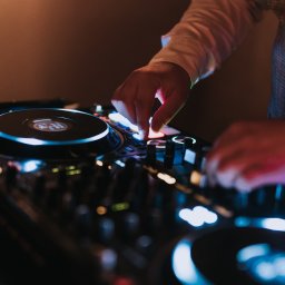 OLSZEWSKI - DJ i Konferansjer - DJ w trakcie występu, miksujący muzykę na profesjonalnym sprzęcie. Widoczne dłonie na kontrolerze, mikrofon w tle i oświetlenie sceniczne.