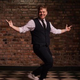 OLSZEWSKI - DJ i Konferansjer - Uśmiechnięty mężczyzna w tańcu, w kamizelce i białych butach, na tle ceglanej ściany i szachownicy na podłodze. Dynamiczna poza, gestykulacja.