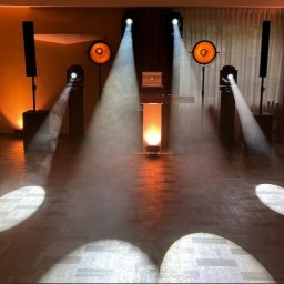 OLSZEWSKI - DJ i Konferansjer - Scena oświetlona reflektorami i lampami, z laptopem na biurku. Pomieszczenie z parkietem, kurtyna w tle. Gotowość do występu.