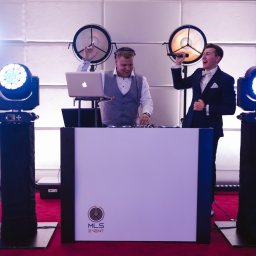 OLSZEWSKI - DJ i Konferansjer - Uśmiechnięty DJ w akcji, obok wokalista w garniturze z mikrofonem. Scena oświetlona reflektorami, nowoczesny sprzęt DJ-ski, dekoracyjne tło.
