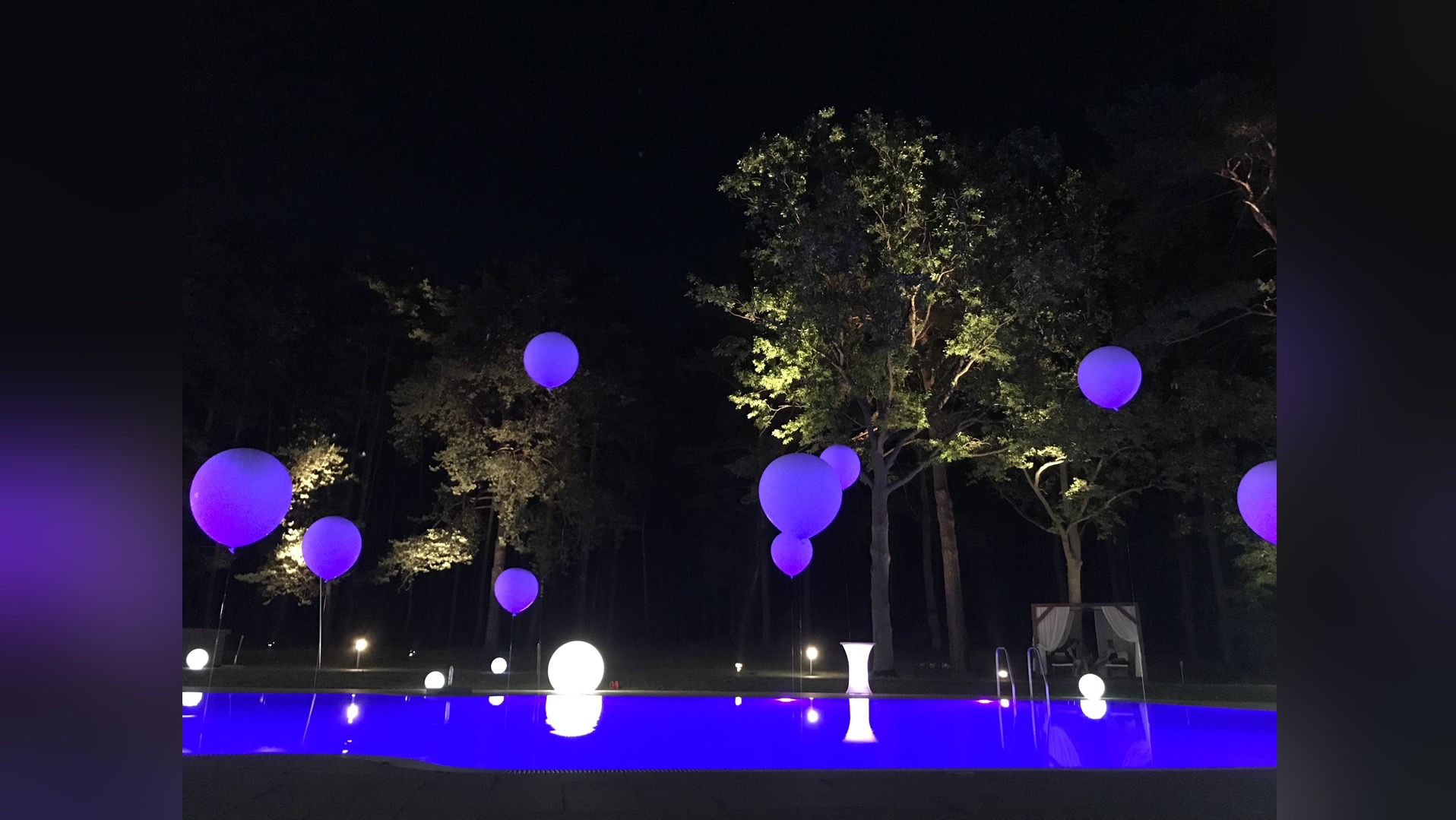 Nocne oświetlenie imprezowe: fioletowe balony i kule nad basenem, drzewa w tle, tworzące magiczną atmosferę.