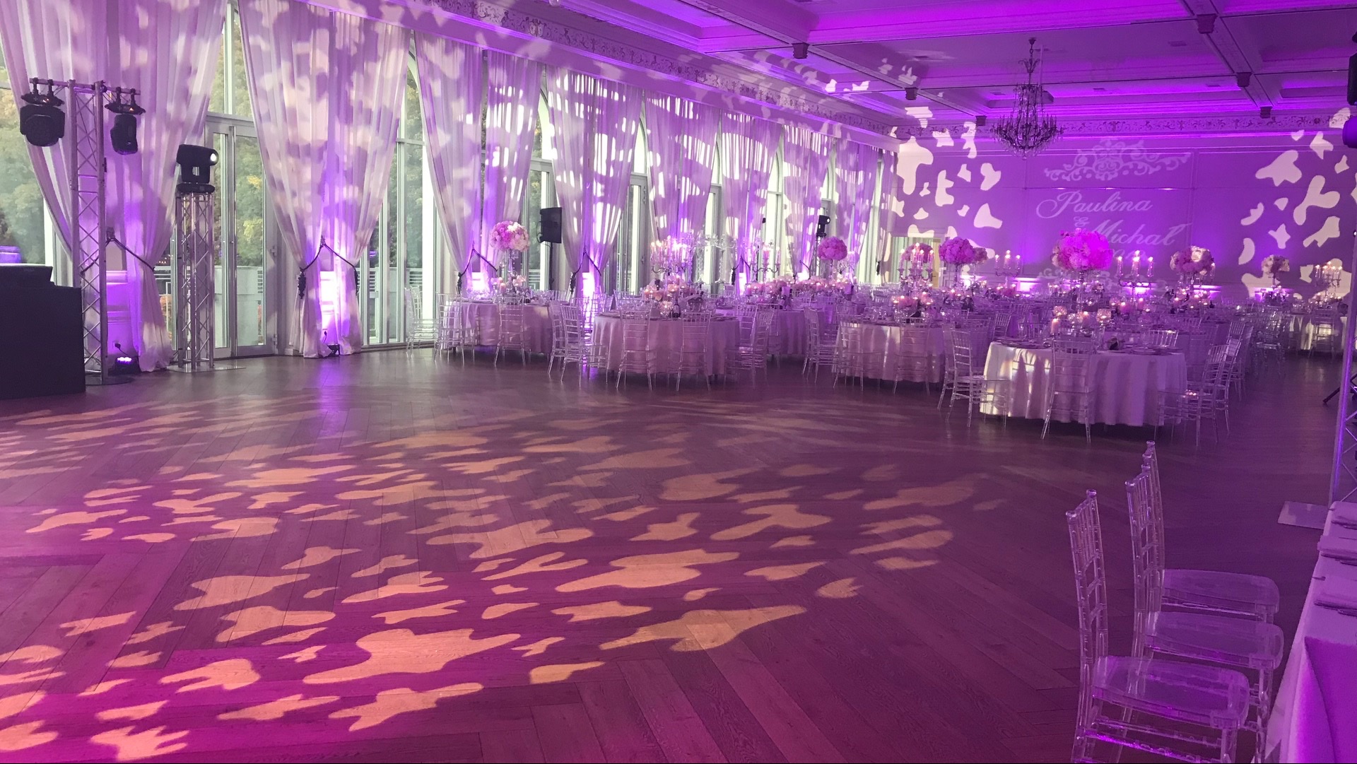 Uroczysta sala balowa z okrągłymi stołami, transparentnymi krzesłami i dekoracjami kwiatowymi, oświetlona fioletowym światłem i wzorami na parkiecie.