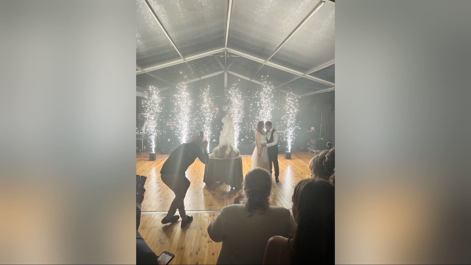 Para młoda kroi tort weselny otoczona fontannami iskier, fotograf robi zdjęcia, widzowie obserwują. Scena w namiocie, drewniana podłoga.