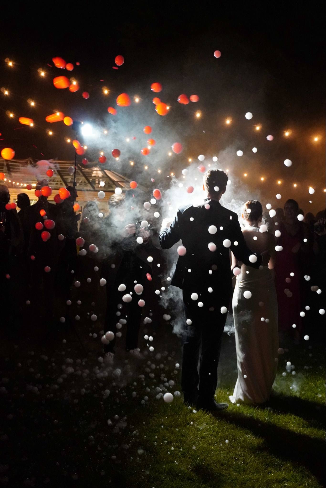 Para młoda w tańcu, otoczona przez mgłę i czerwono-białe bańki, pod girlandami świateł podczas weselnego przyjęcia w plenerze, widok z tyłu.