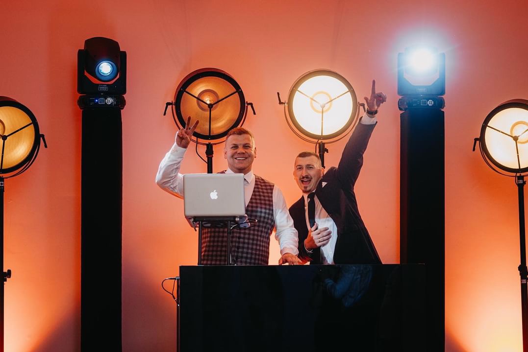Uśmiechnięci muzycy za konsoletą DJ-ską, z laptopem Apple i profesjonalnym oświetleniem estradowym w tle, w trakcie występu.
