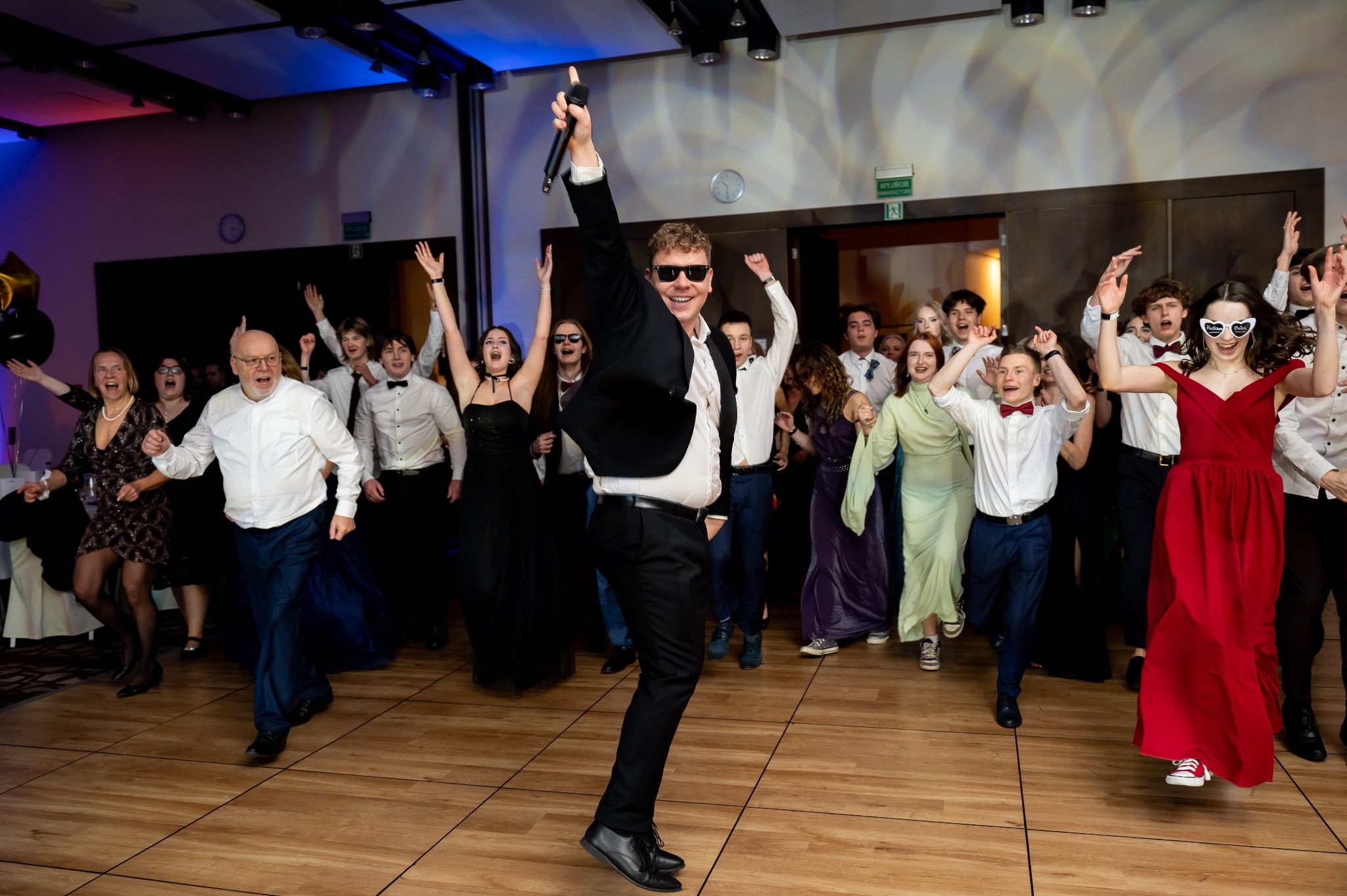 Rozbawiony wokalista w okularach przeciwsłonecznych, z mikrofonem w dłoni, w otoczeniu tańczącego tłumu na parkiecie. Uczestnicy w strojach wieczorowych.